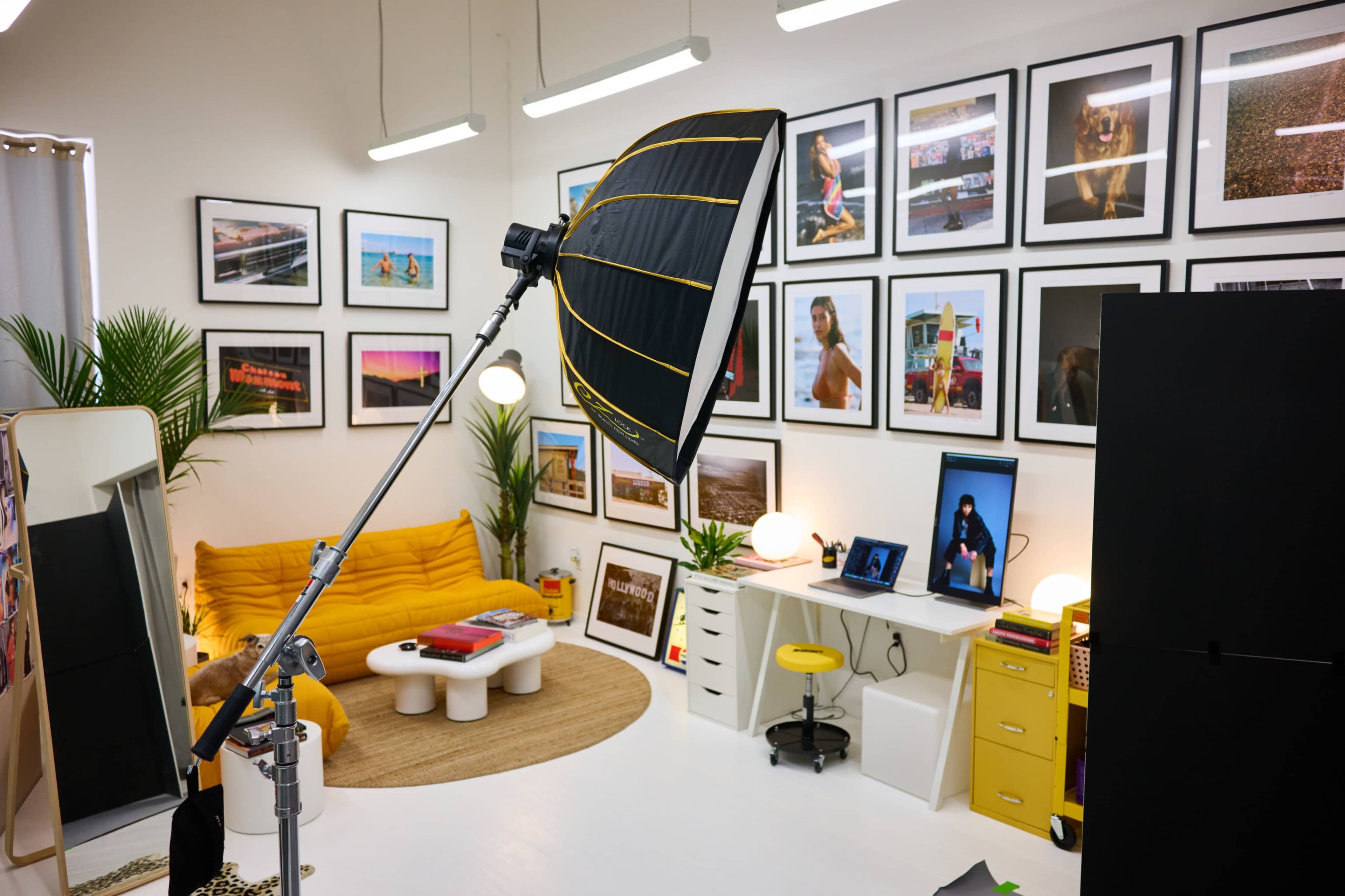 A brightly lit studio space featuring a large umbrella light, a yellow sofa, a variety of framed photographs on the walls, and a workspace with a computer and yellow accents.