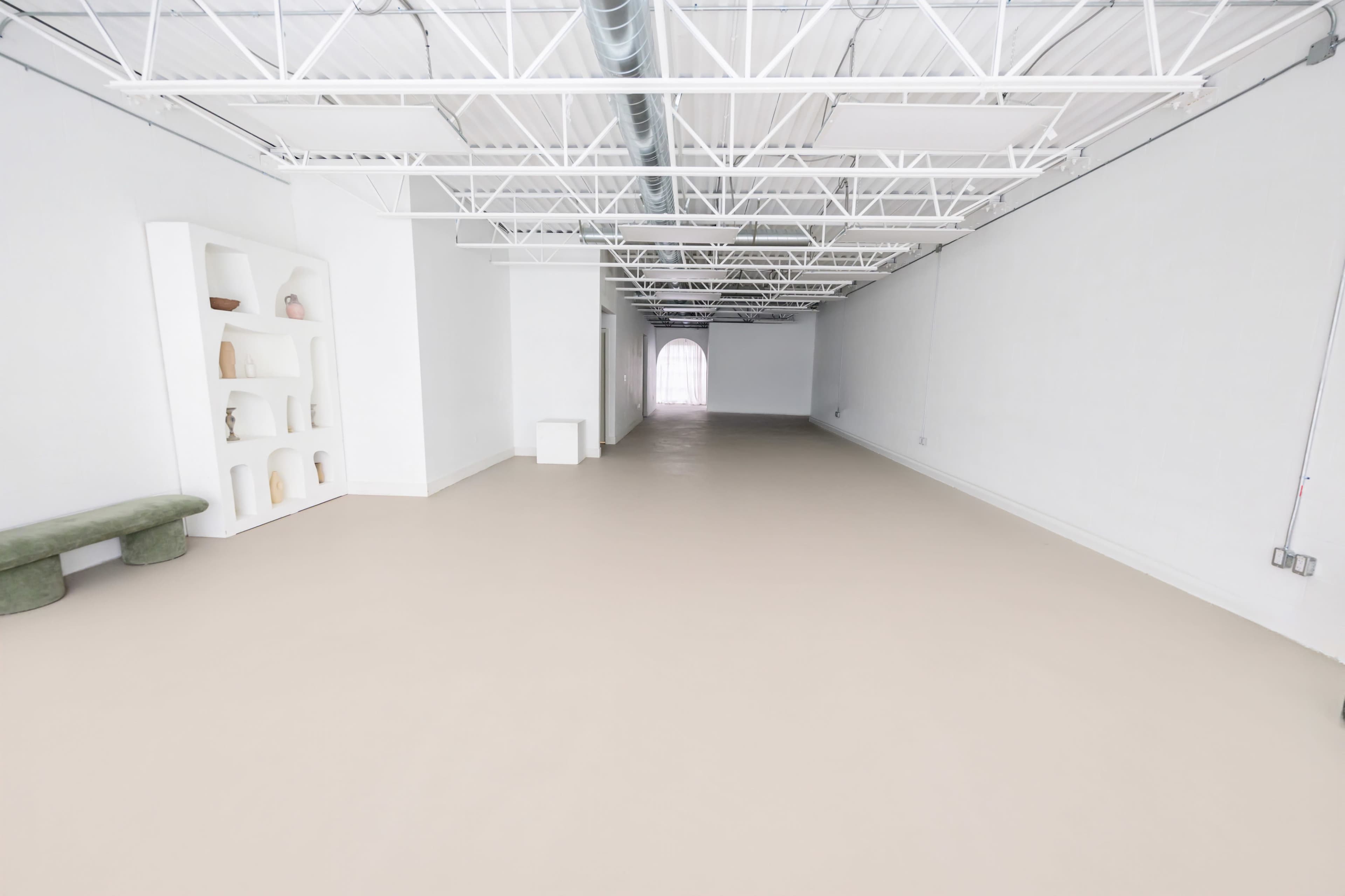 The image shows a spacious, empty interior with white walls, a light-colored floor, and a minimalist display shelf on the left.