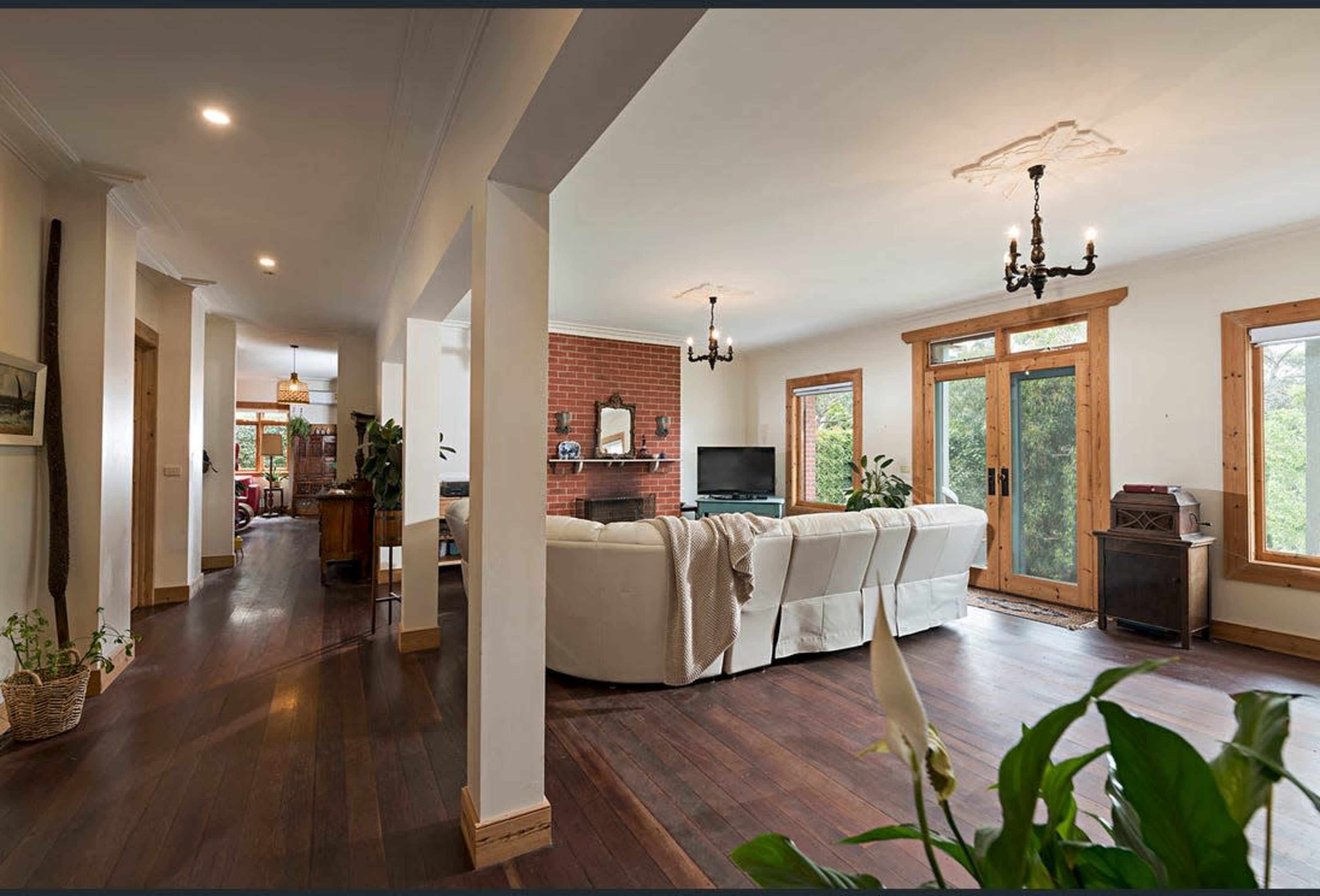 The image shows a spacious living room with wooden floors, large windows, and a white sectional sofa facing a brick fireplace and a television.