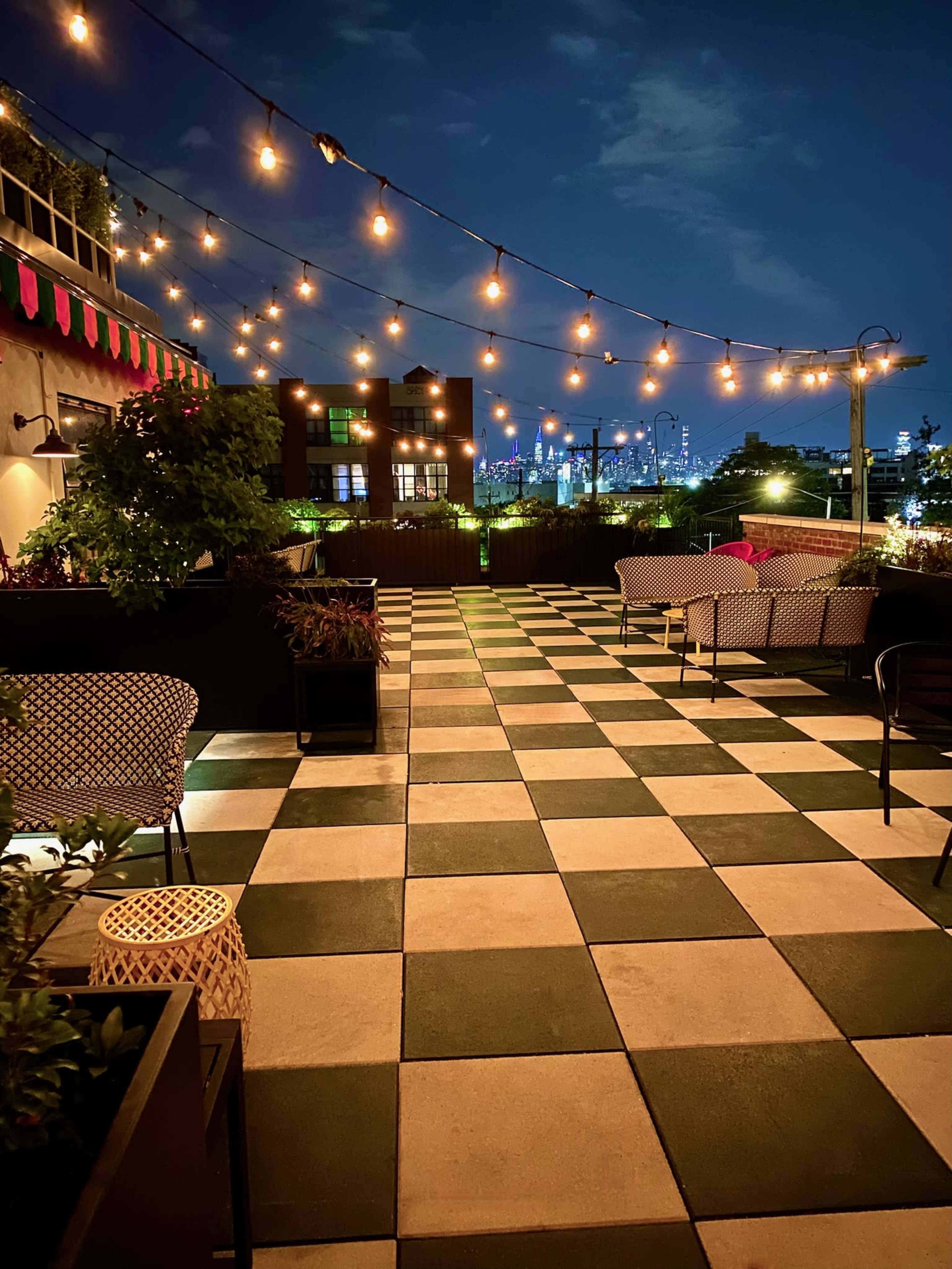 The image shows a rooftop patio at night, featuring checkered flooring, string lights overhead, and a view of city buildings in the background.