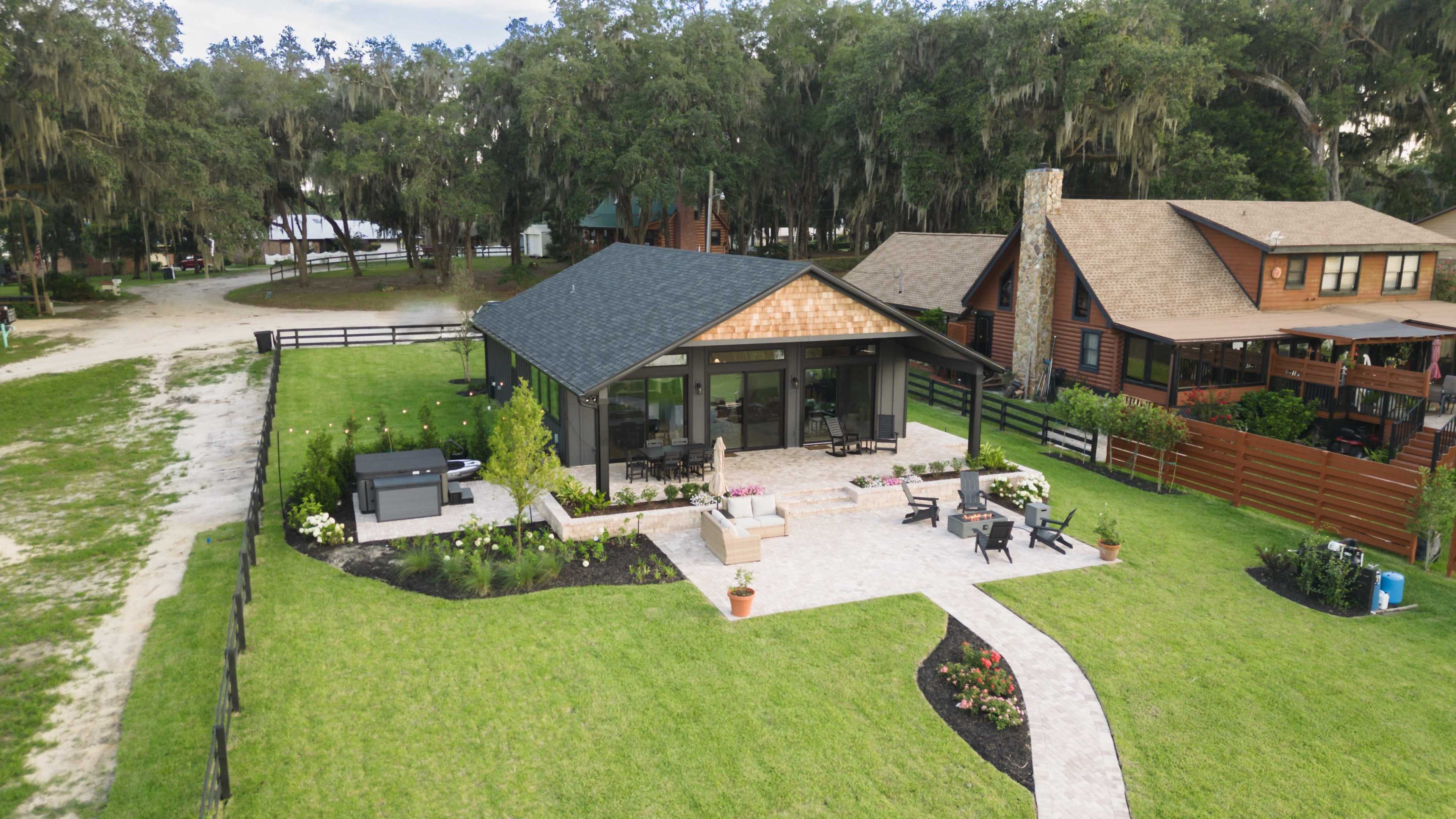 A modern house surrounded by landscaped lawn, paved pathways, and seating areas, situated near wooded areas and additional cabins.