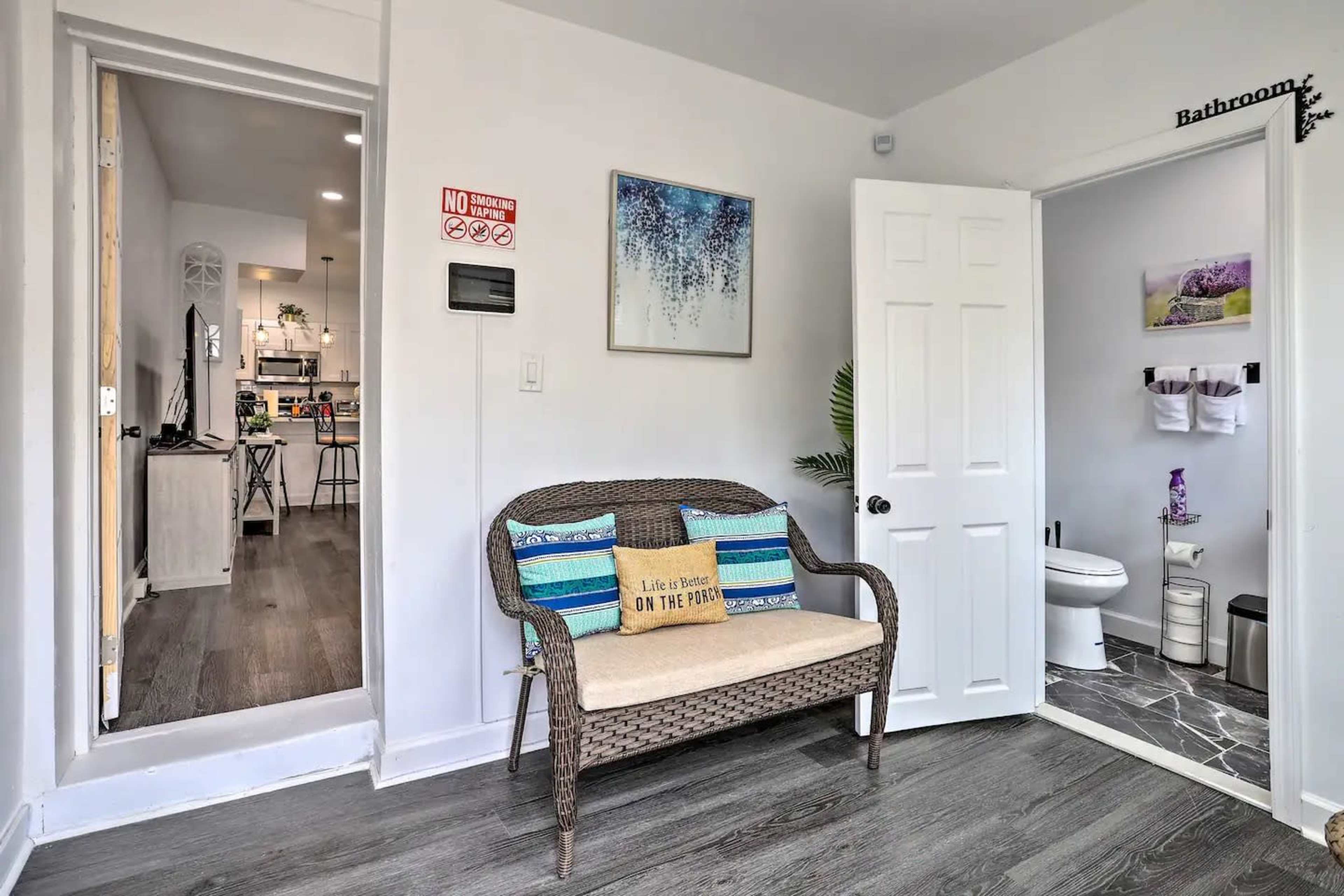 The image shows a small entryway with a wicker bench, a door leading to a bathroom, and a view into a kitchen area.
