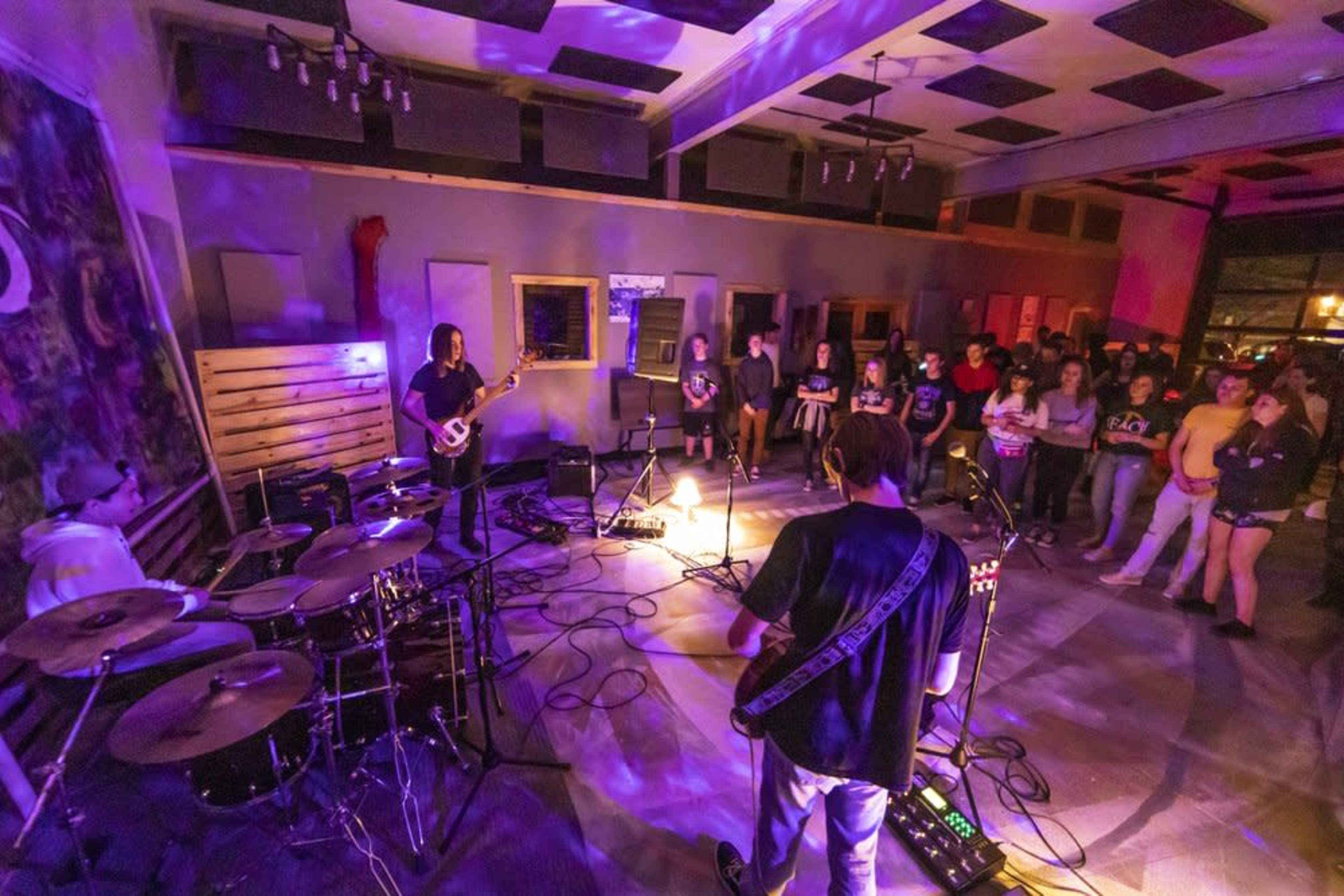 A band performs on a low-lit stage while an audience watches intently in a venue filled with purple lighting.
