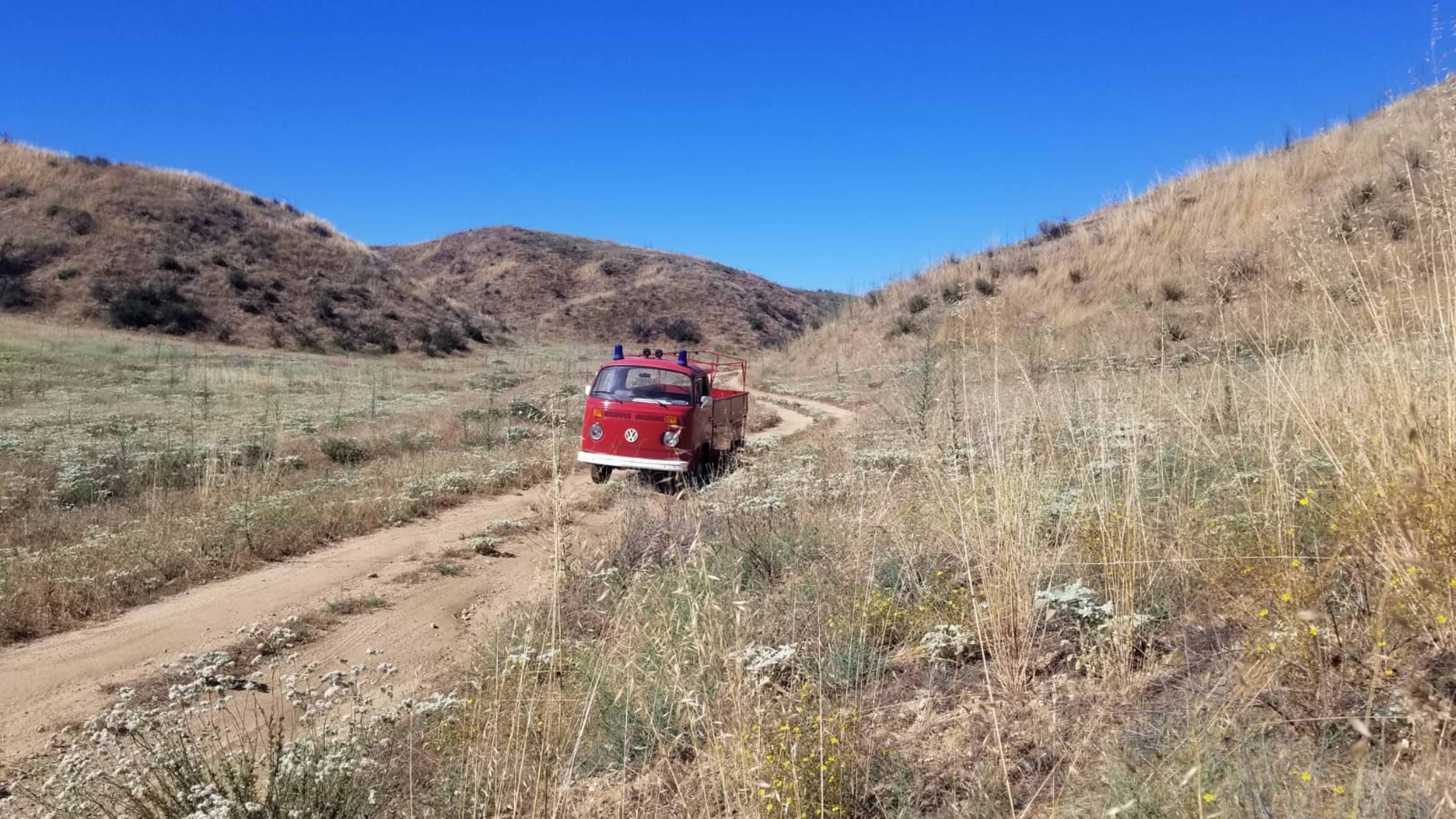 A red vintage fire truck drives along a dirt path amidst a grassy landscape with rolling hills.