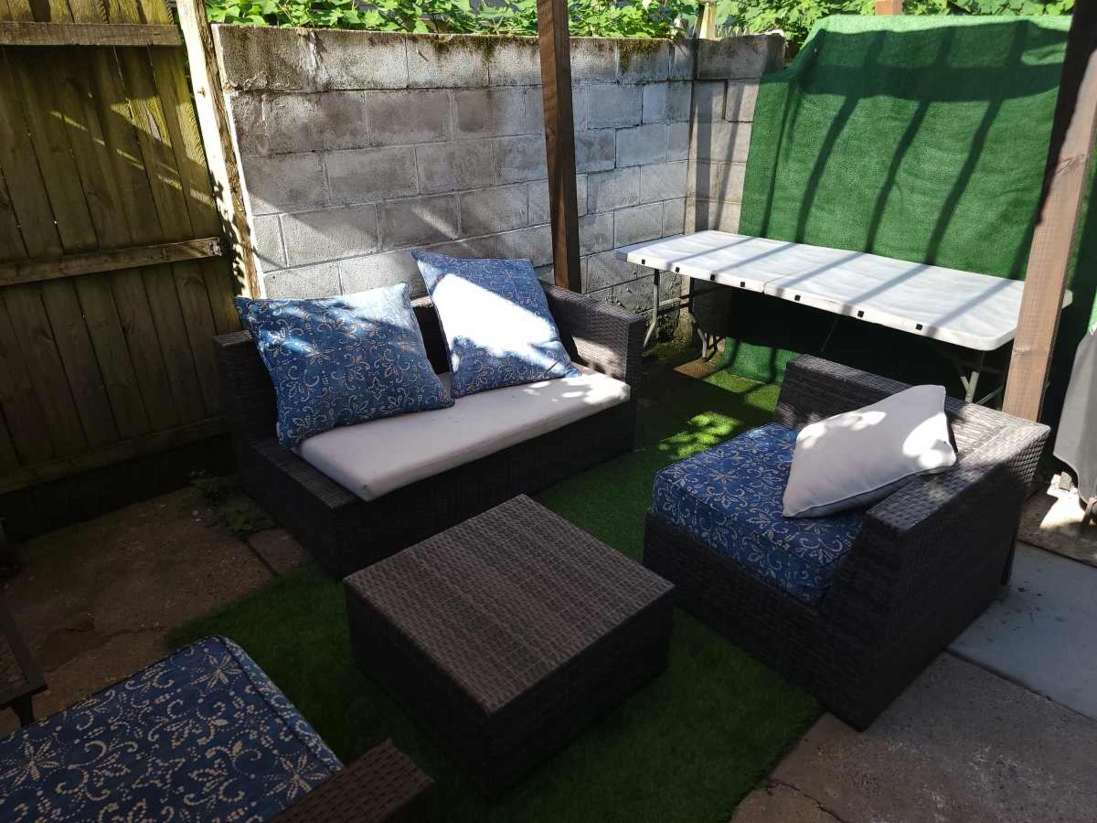 The scene shows a small outdoor seating area with wicker furniture and blue-patterned cushions, surrounded by a wooden fence and a green tarp.