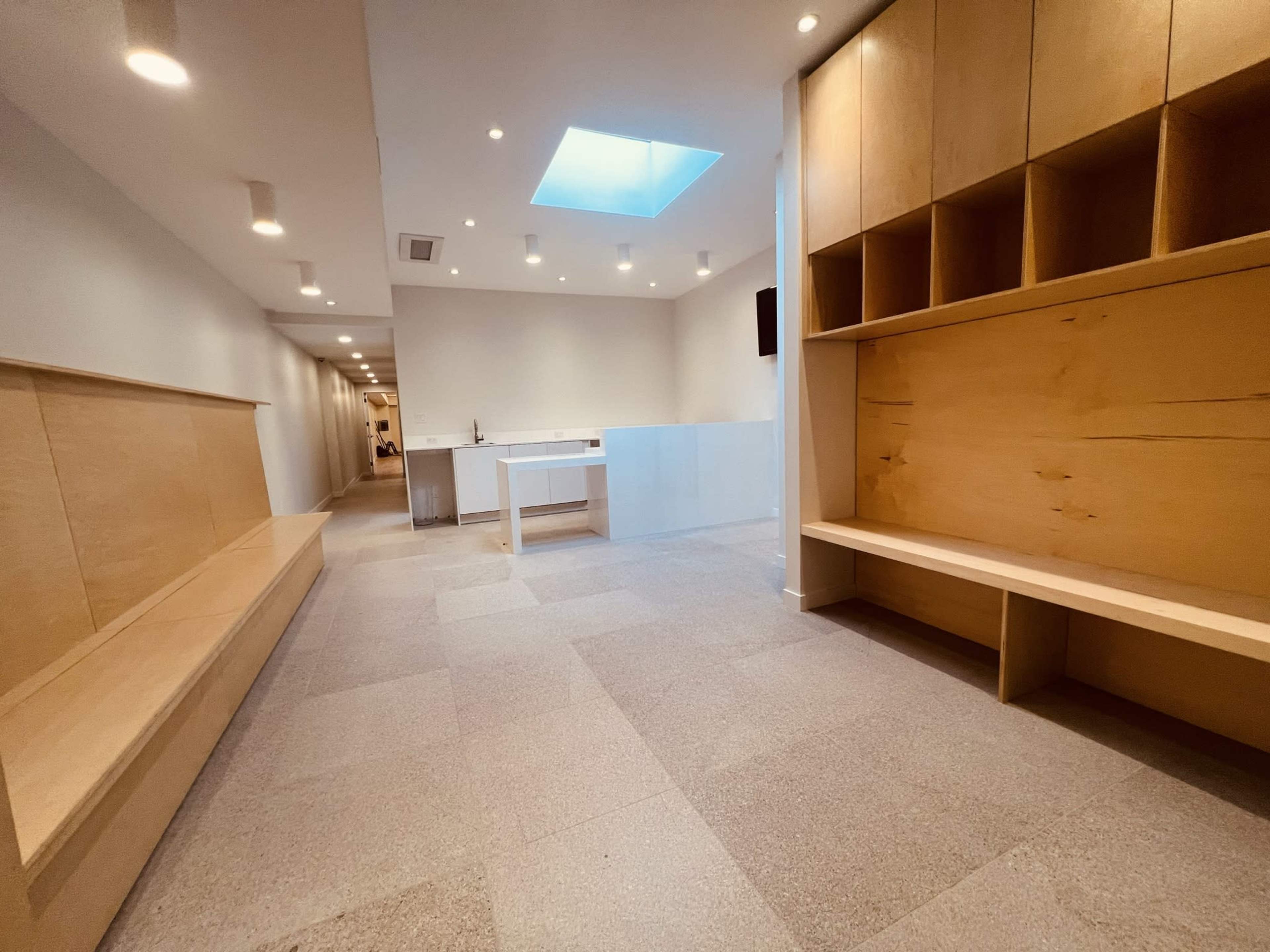 A minimalist interior space with light-colored wood panels, a skylight, and a stone floor, featuring benches along one wall and a white table in the center.