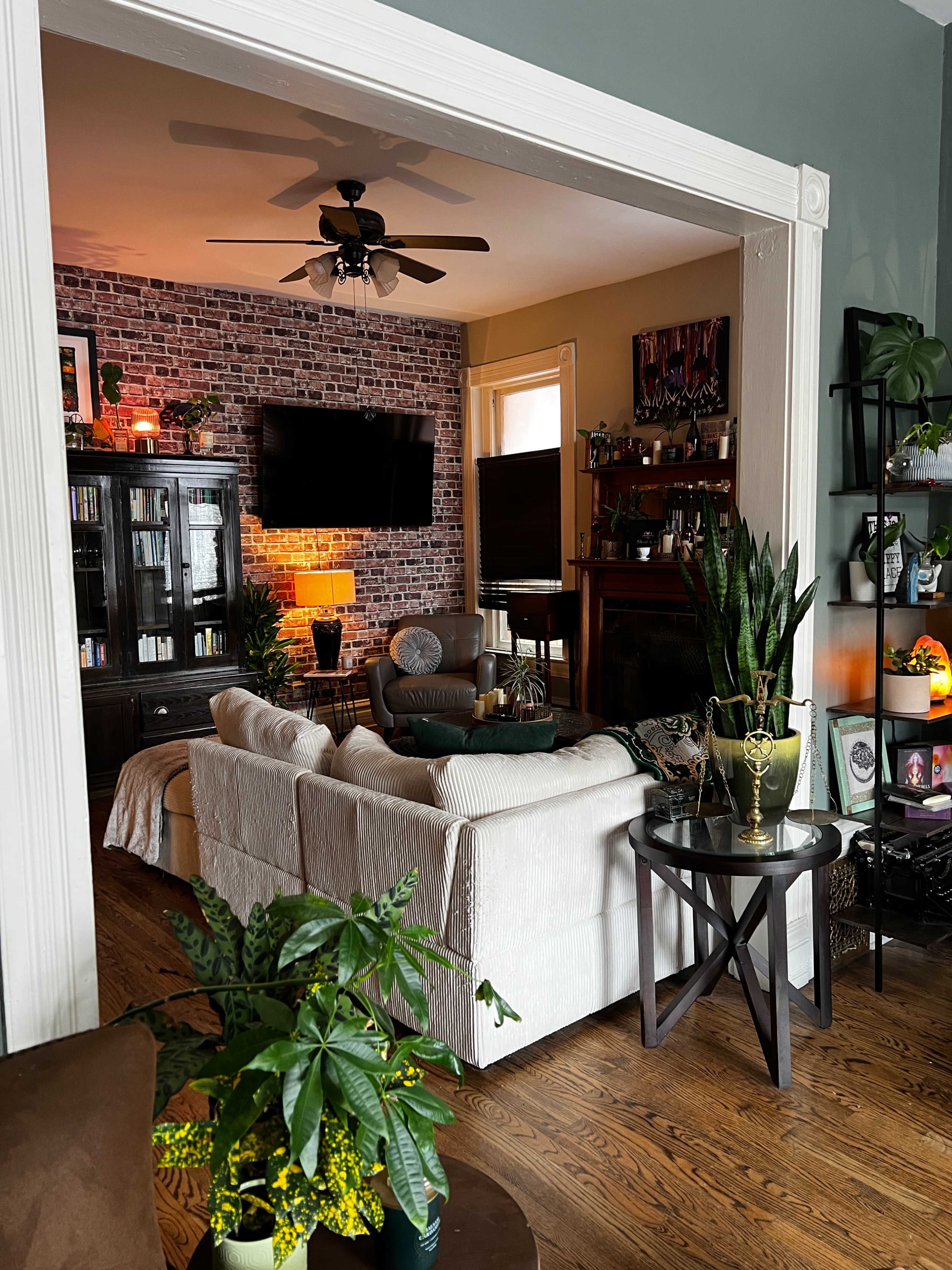 A cozy living room features a brick accent wall, a sectional sofa, a large television, and plants decorating the space.