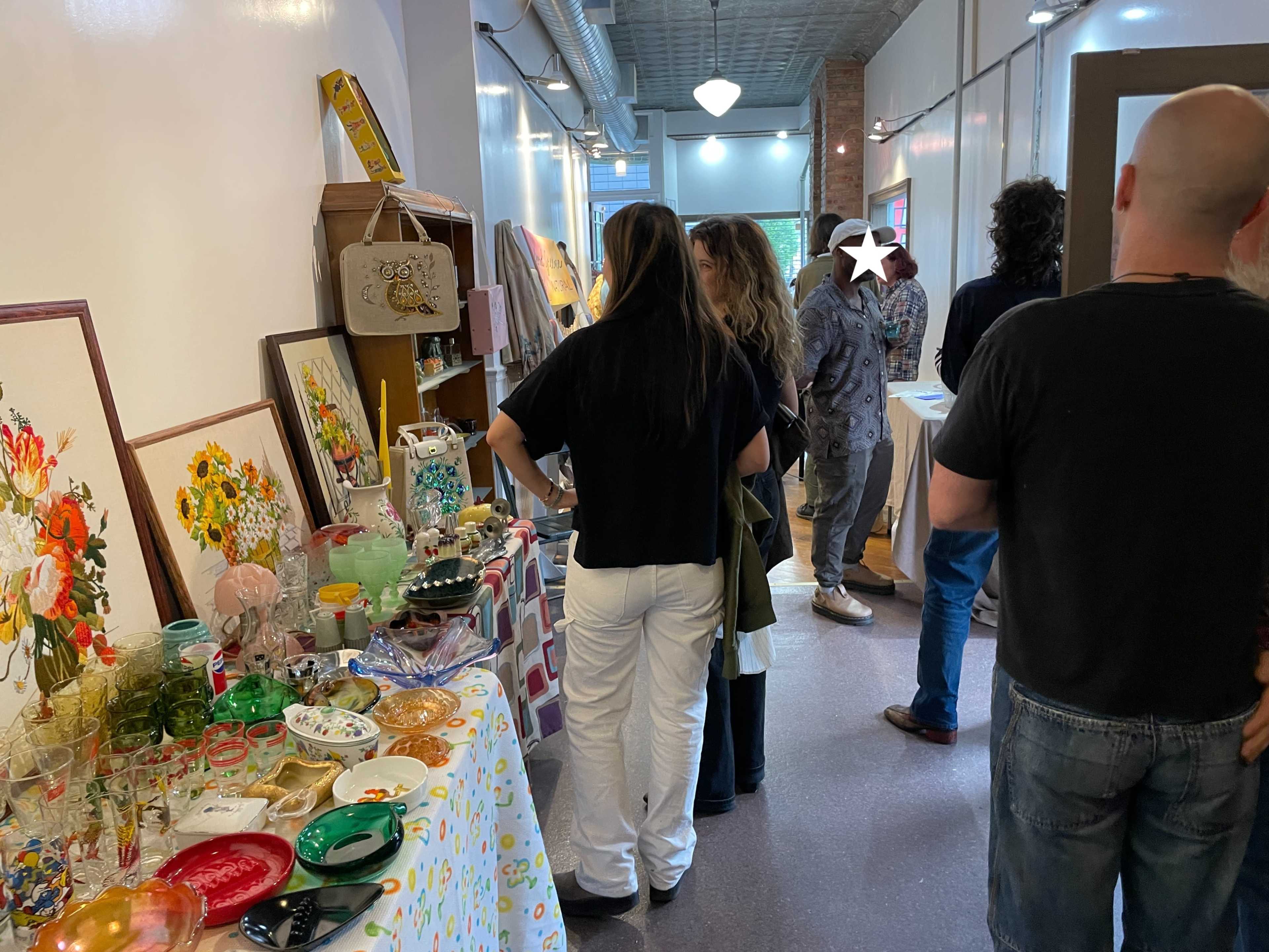A group of people browses through displayed items, including colorful dishes and artwork, in a well-lit indoor space.