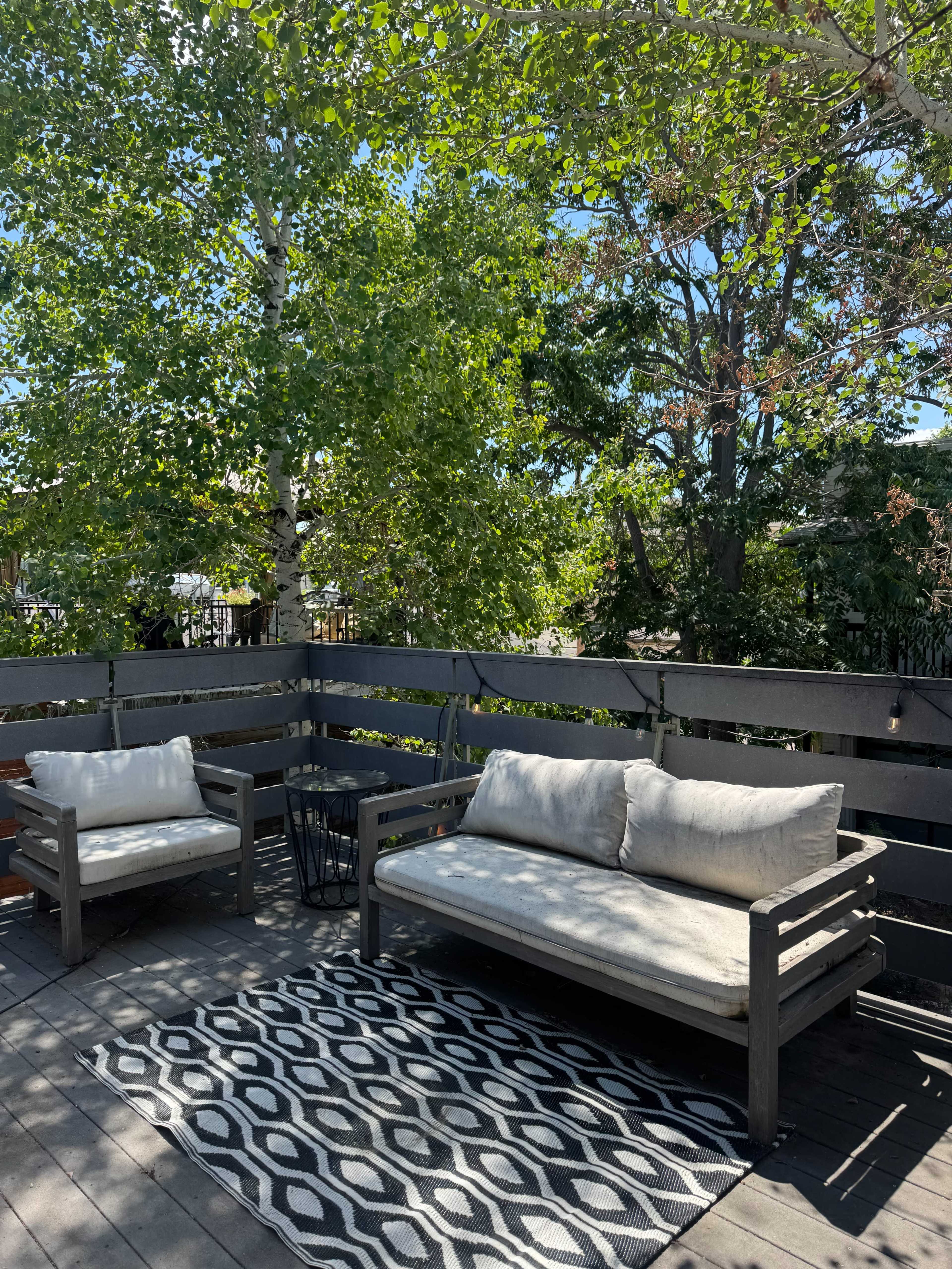 A patio area features two wooden sofas with cushions, a black and white patterned rug, and surrounded by green trees.