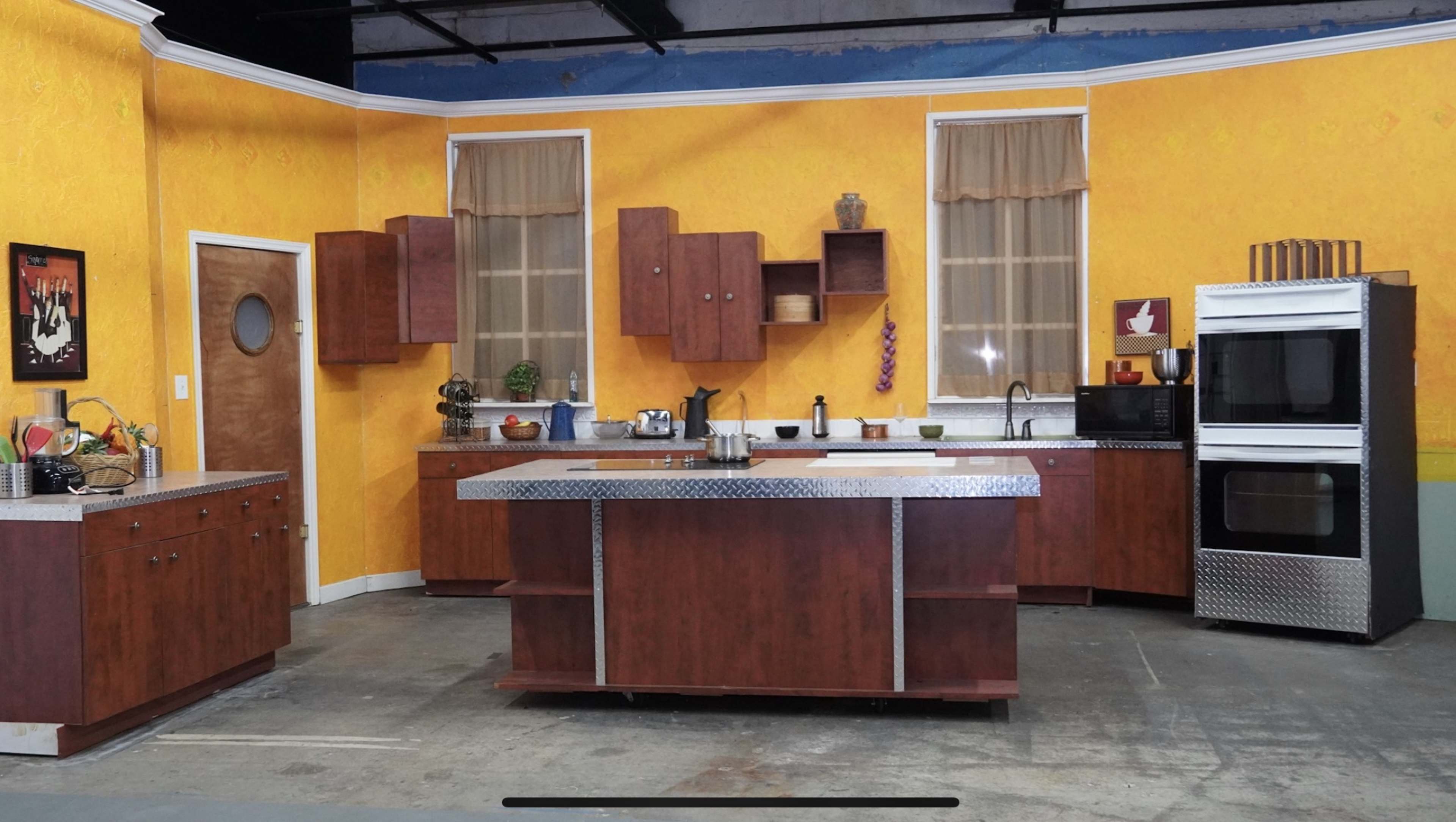 The image shows a kitchen set with yellow walls, wooden cabinets, and stainless steel appliances.