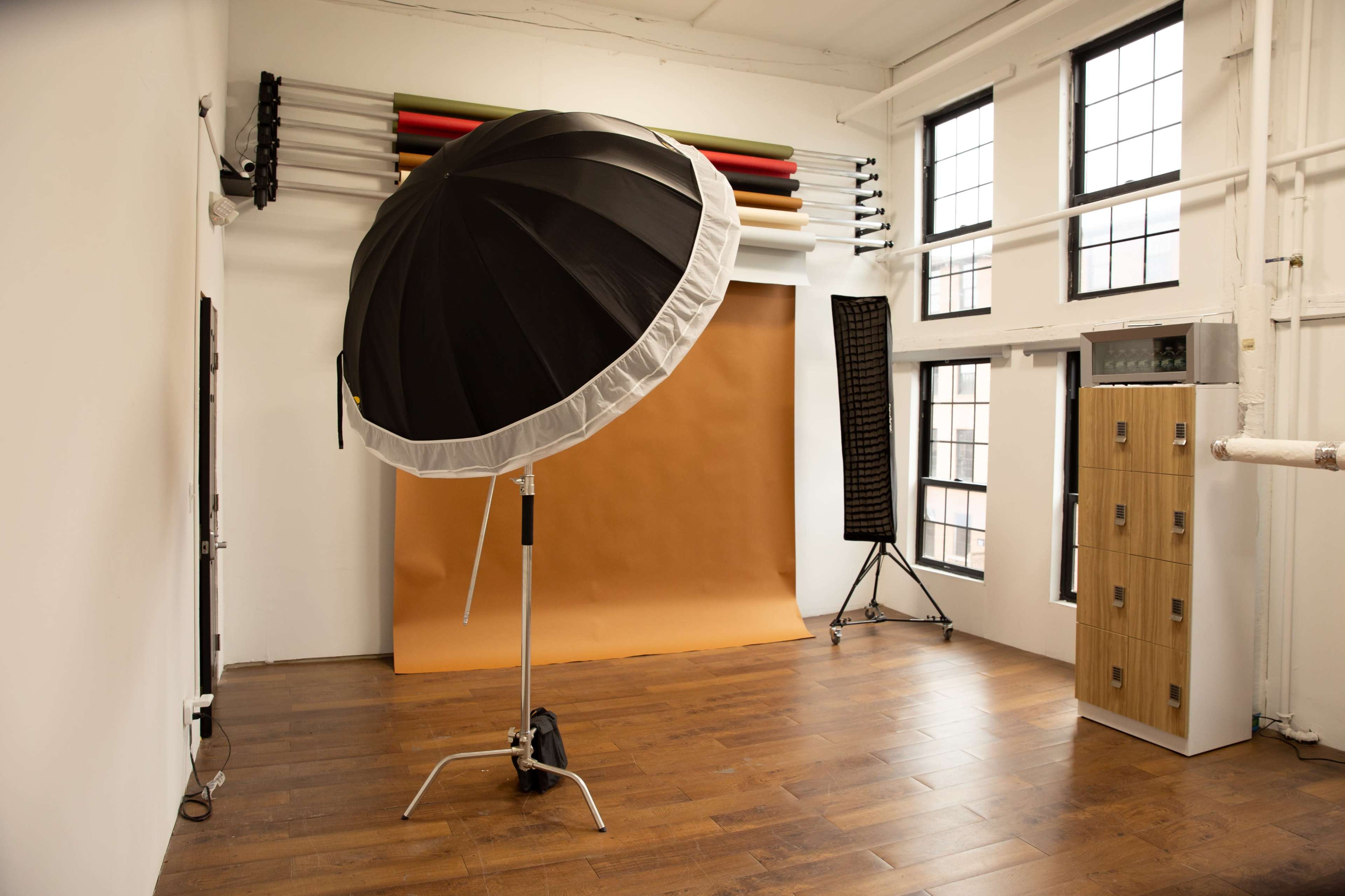 The image shows a photography studio with a large black umbrella softbox, a backdrop of brown paper, and several windows letting in natural light.
