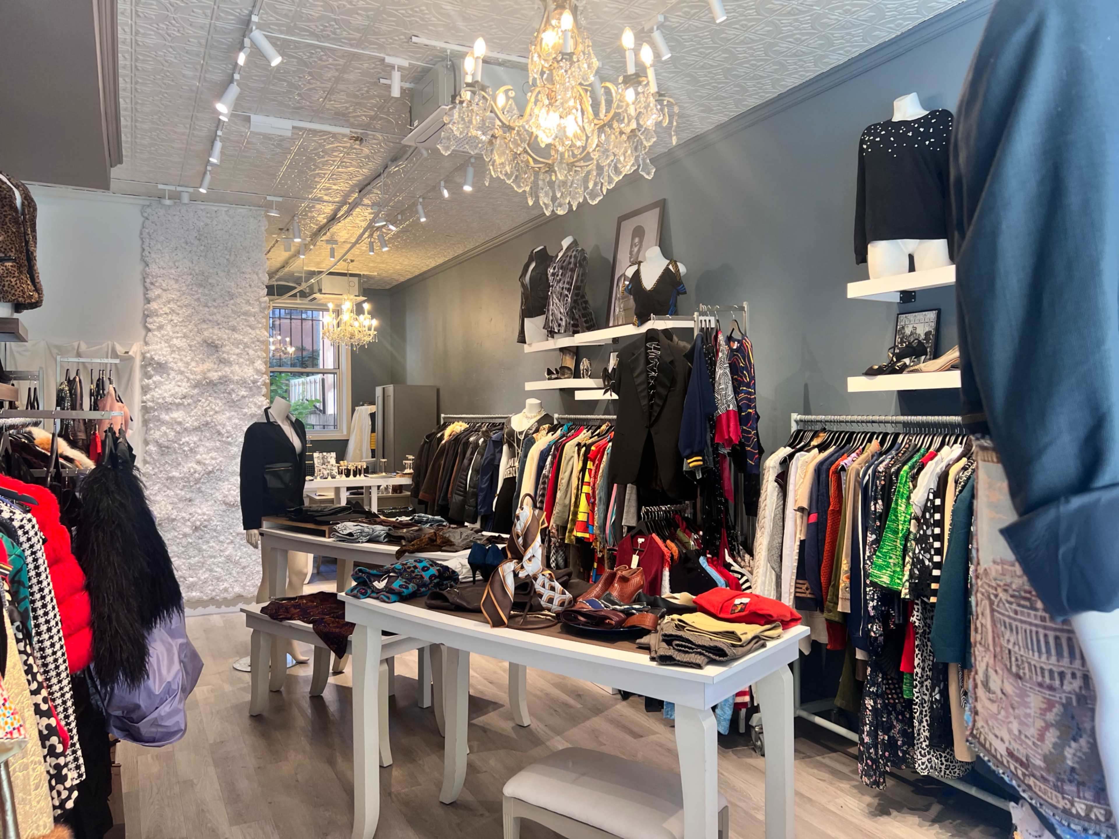 The image shows a clothing boutique featuring rows of apparel on racks, tables displaying accessories, and decorative lighting fixtures.