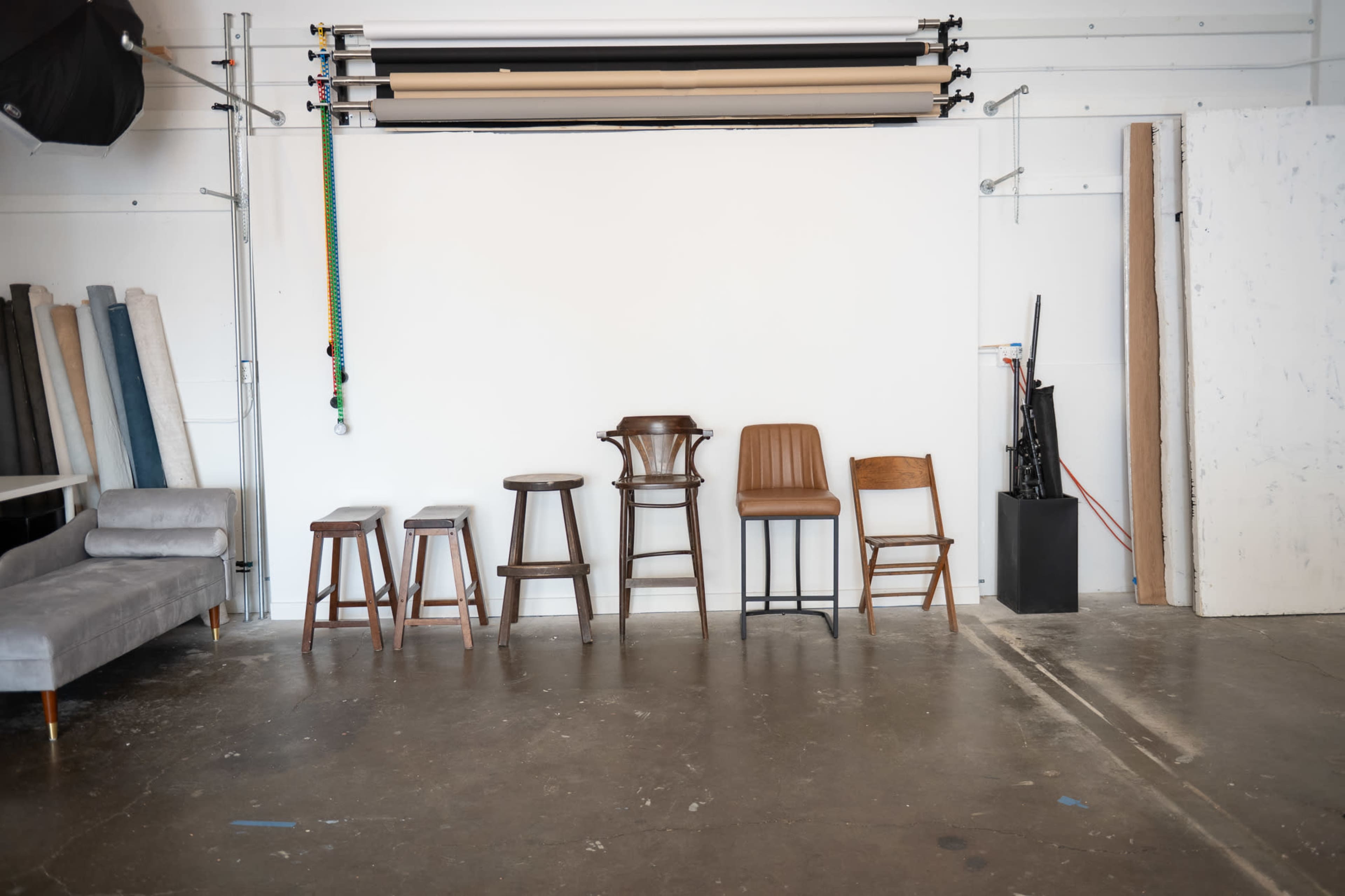 The image shows a row of six different wooden chairs lined up against a plain white wall in a studio space, with various backdrop materials stacked to one side.