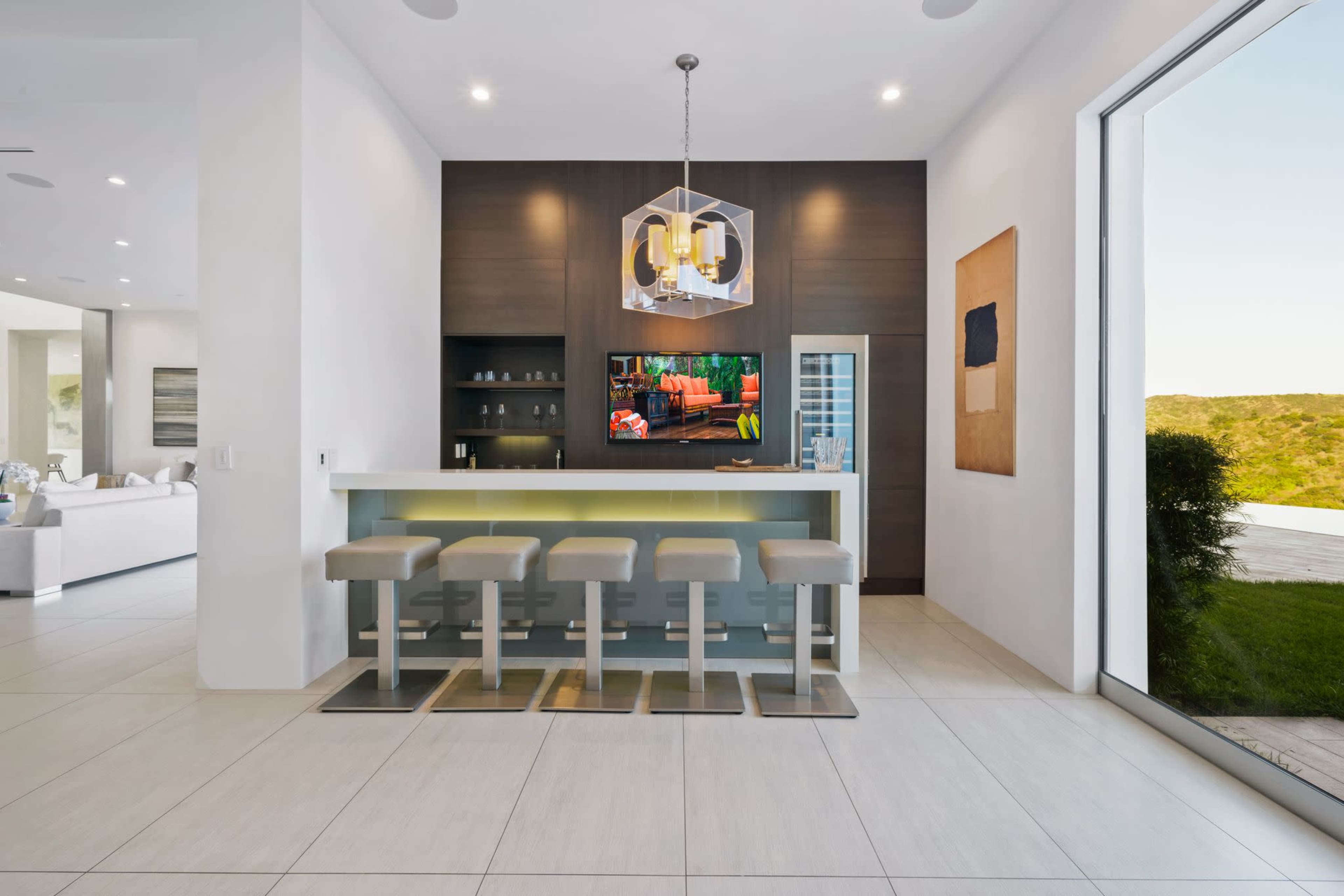 A modern kitchen bar with four stools, a television mounted on the wall, and large windows overlooking a green landscape.
