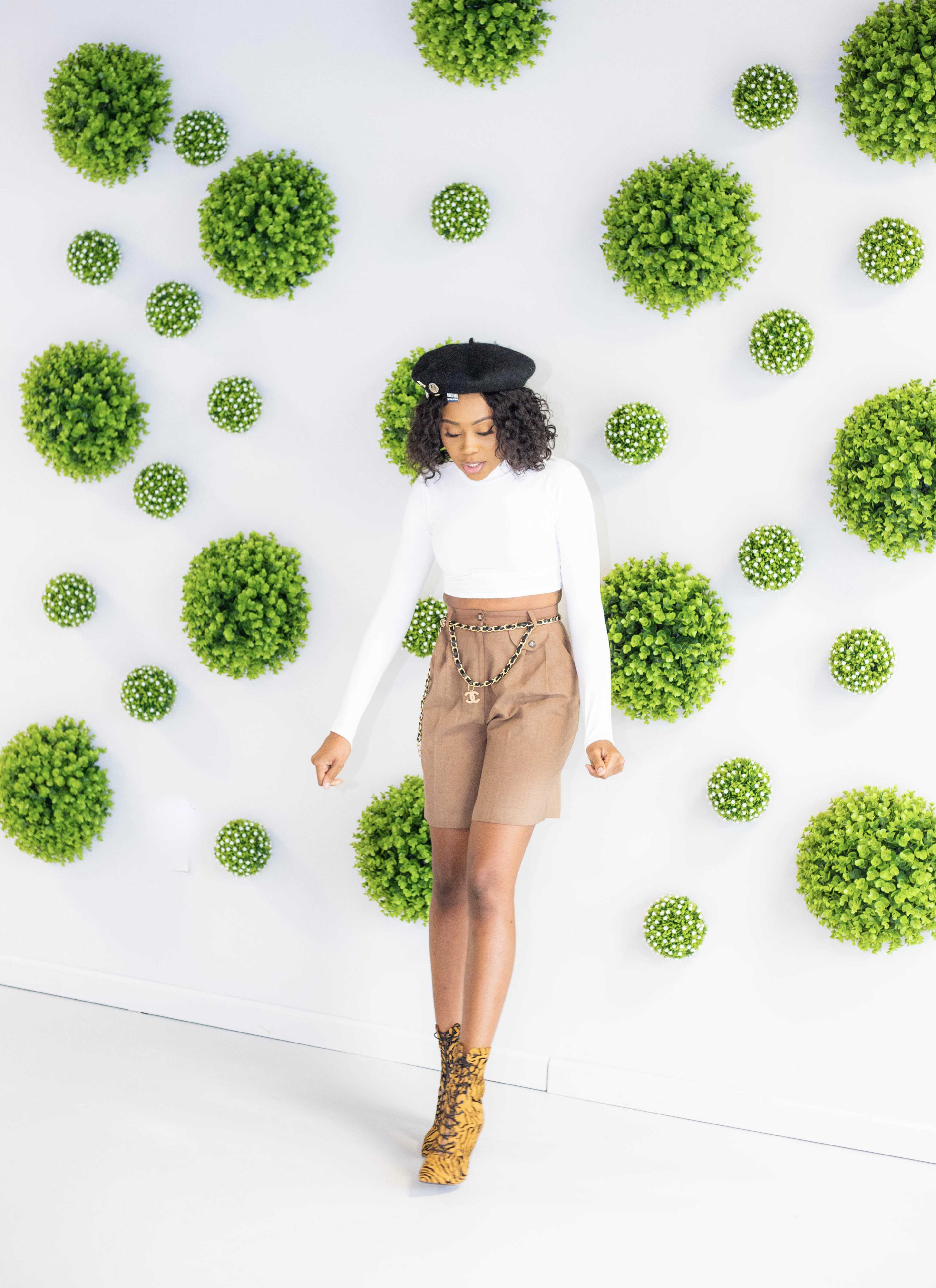 A person in a white long-sleeve top and brown skirt stands against a wall adorned with spherical green plants.