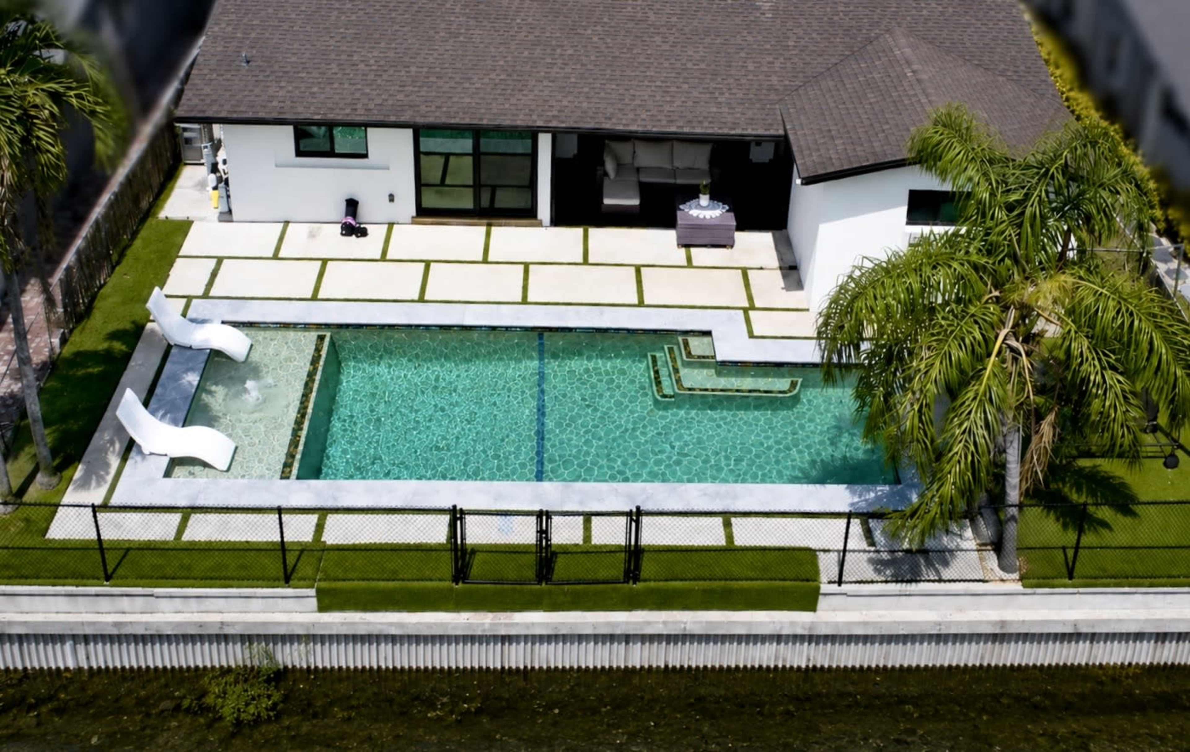 The image shows a modern house adjacent to a rectangular swimming pool surrounded by neatly arranged tiles and green landscaping.