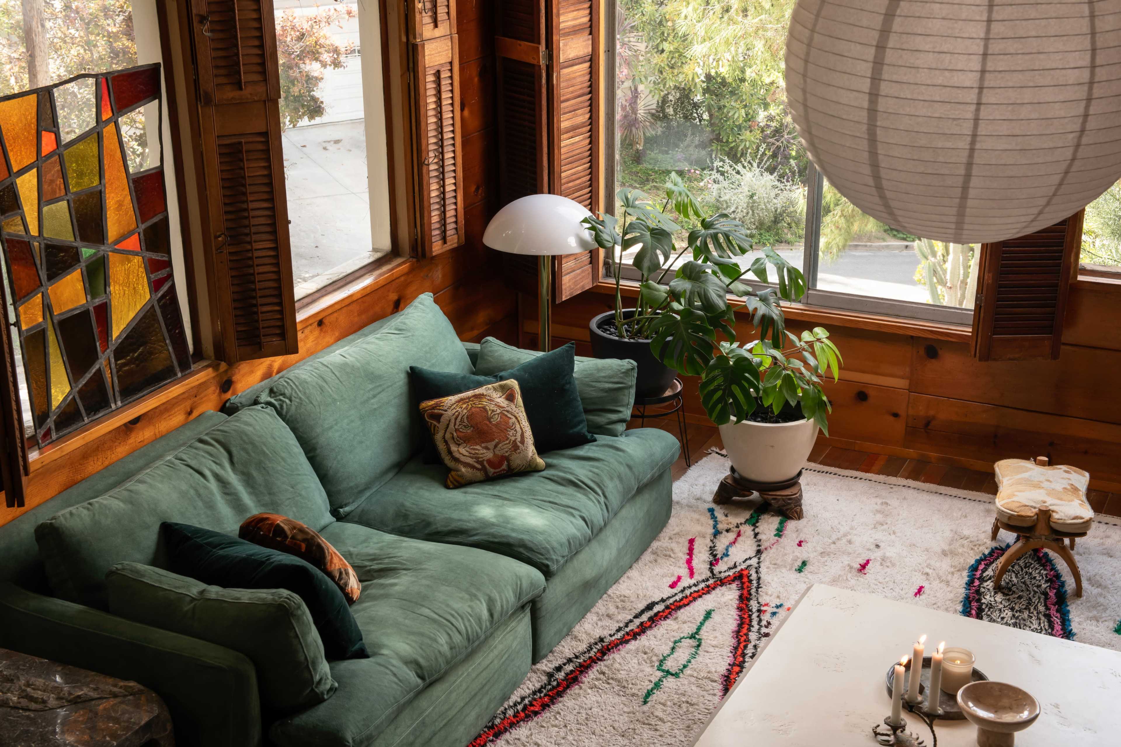 A cozy living room with a green sectional sofa, a large window letting in natural light, and a potted plant near a coffee table on a patterned rug.