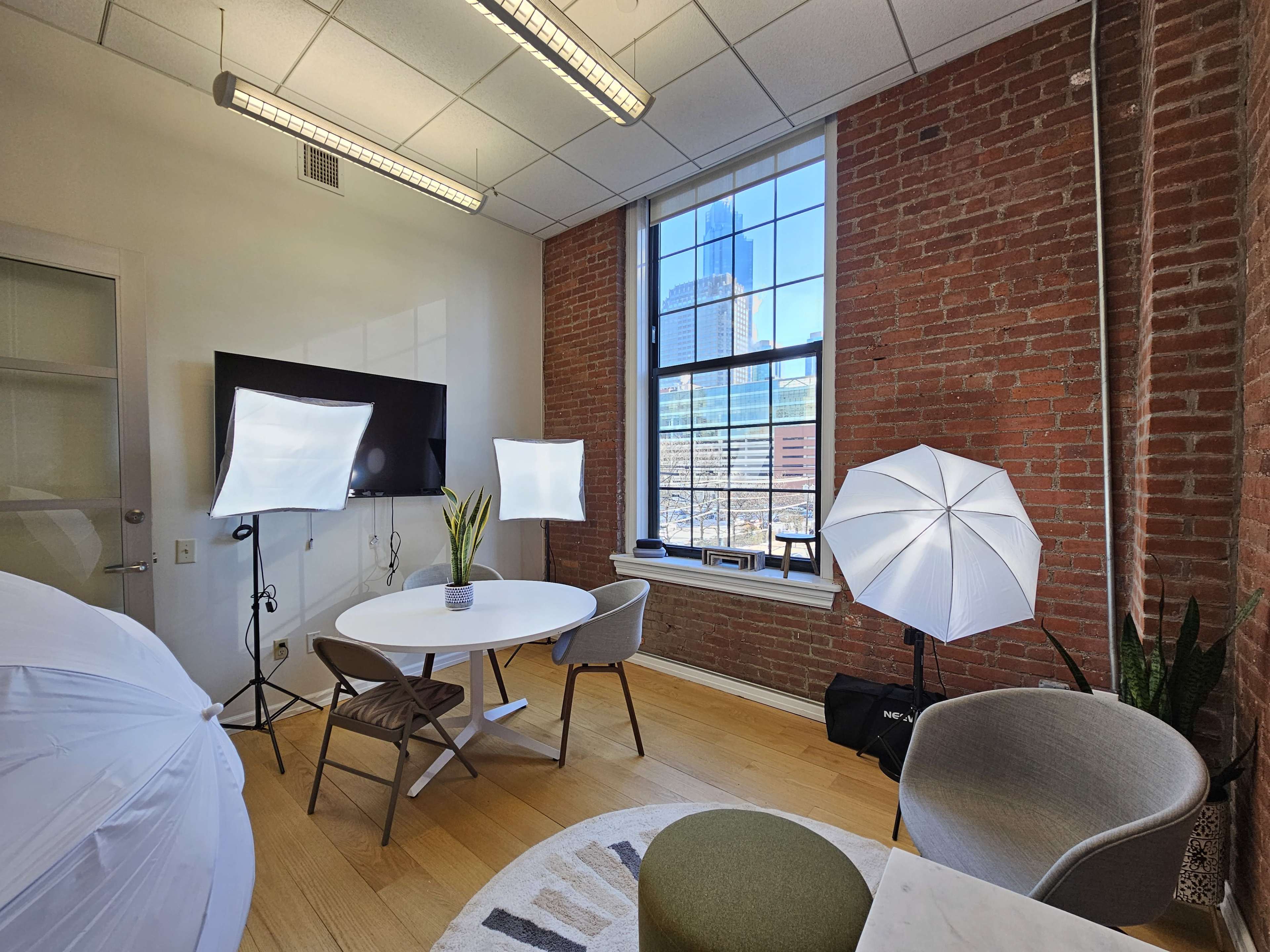 The image shows a photography studio with a round table, chairs, lighting equipment, and large windows displaying a cityscape.
