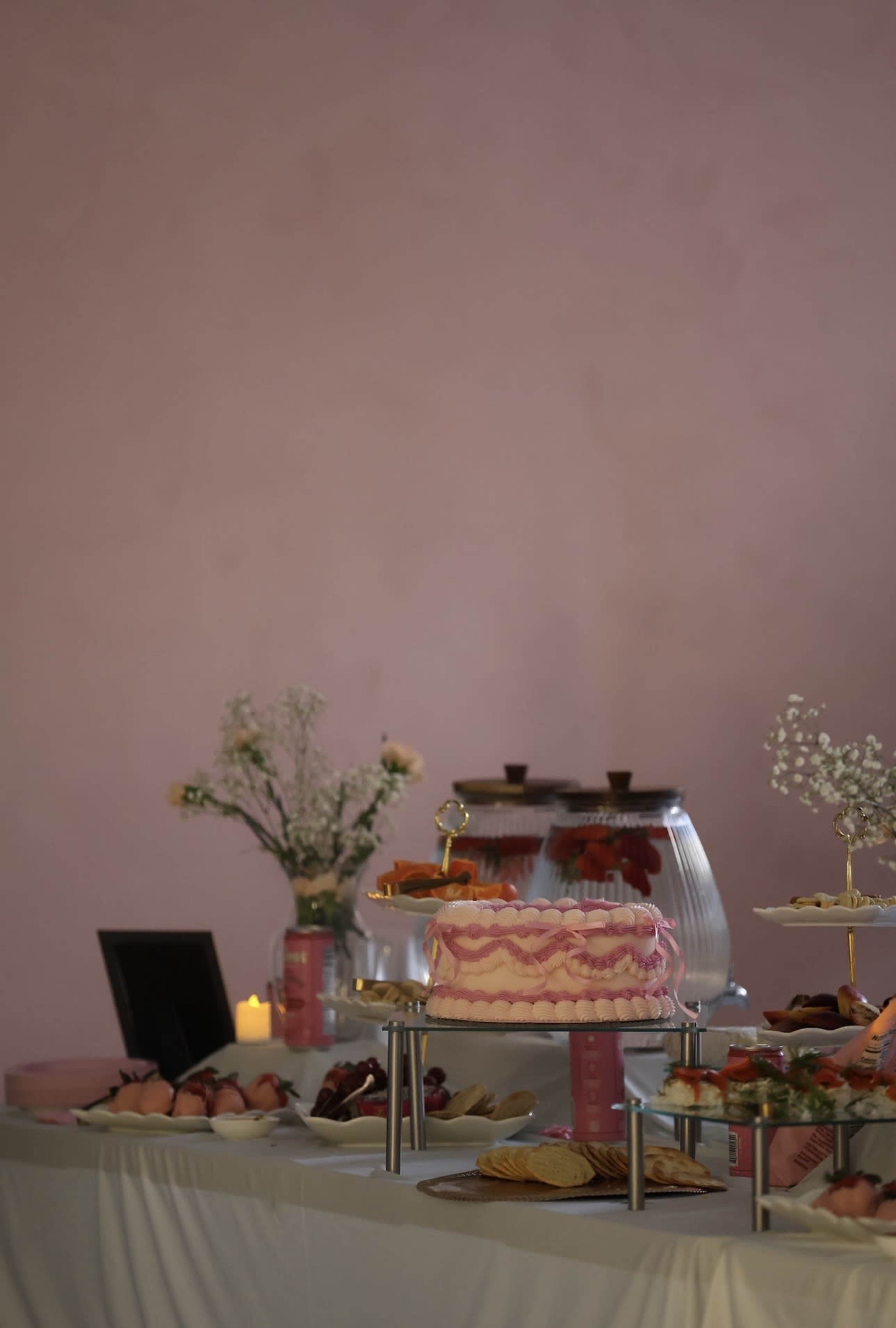 A pink cake sits on a stand among an array of desserts and floral arrangements on a white tablecloth against a pink wall.