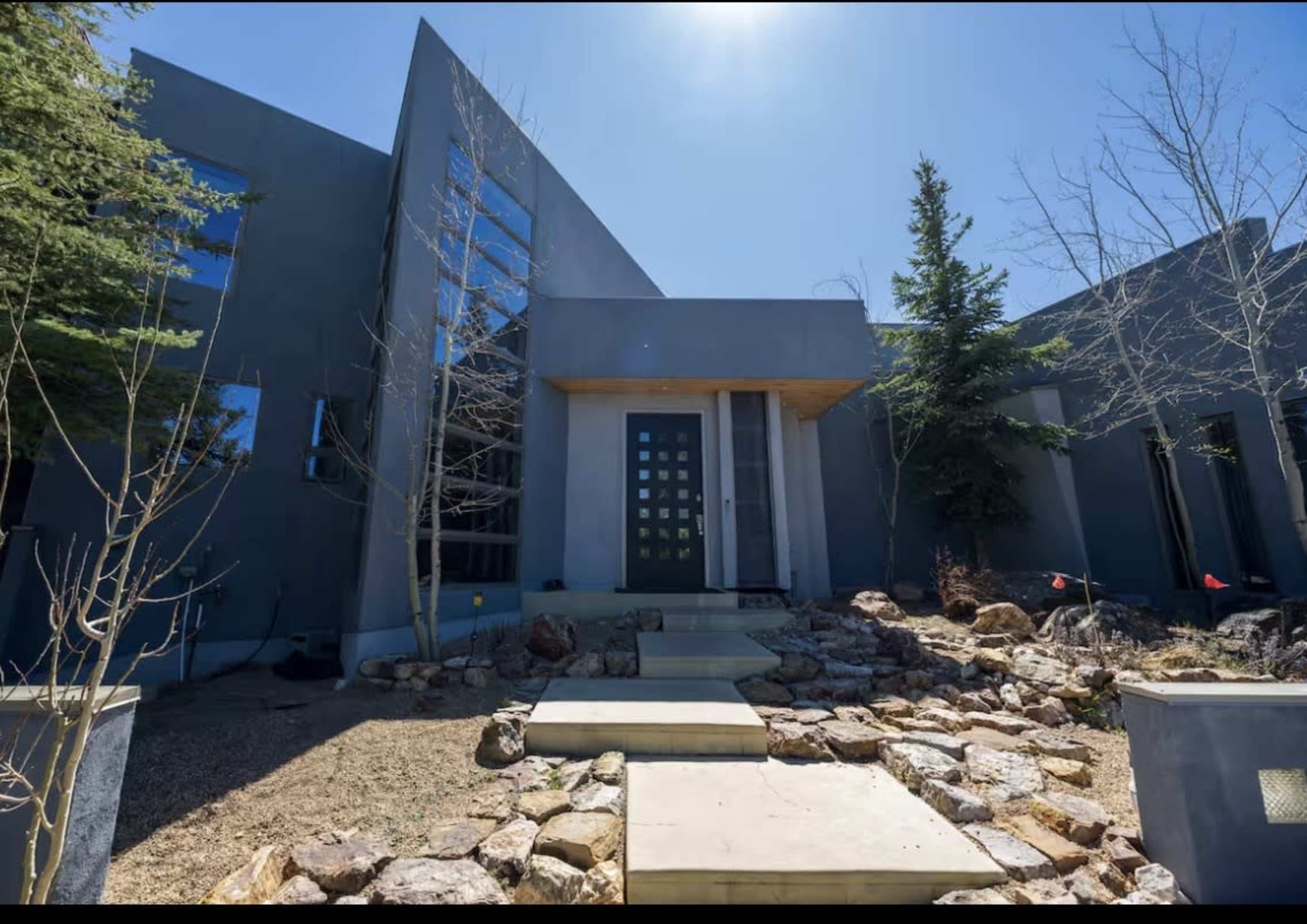 The image shows a modern, angular house with large windows, a stone path leading to the entrance, and sparse landscaping featuring rocks and a few trees.