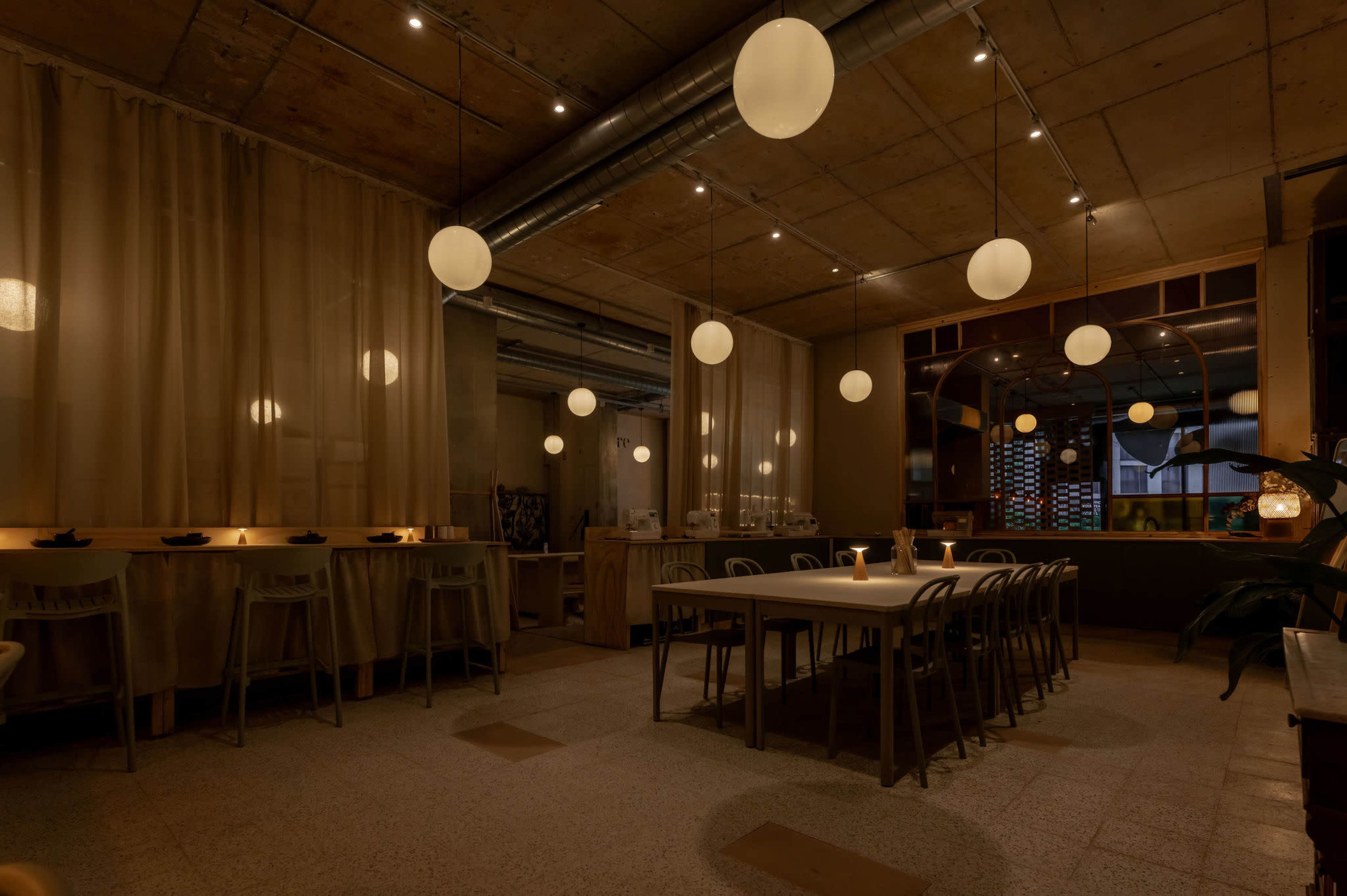 A dimly lit interior of a modern restaurant features long tables and round lanterns hanging from the ceiling.