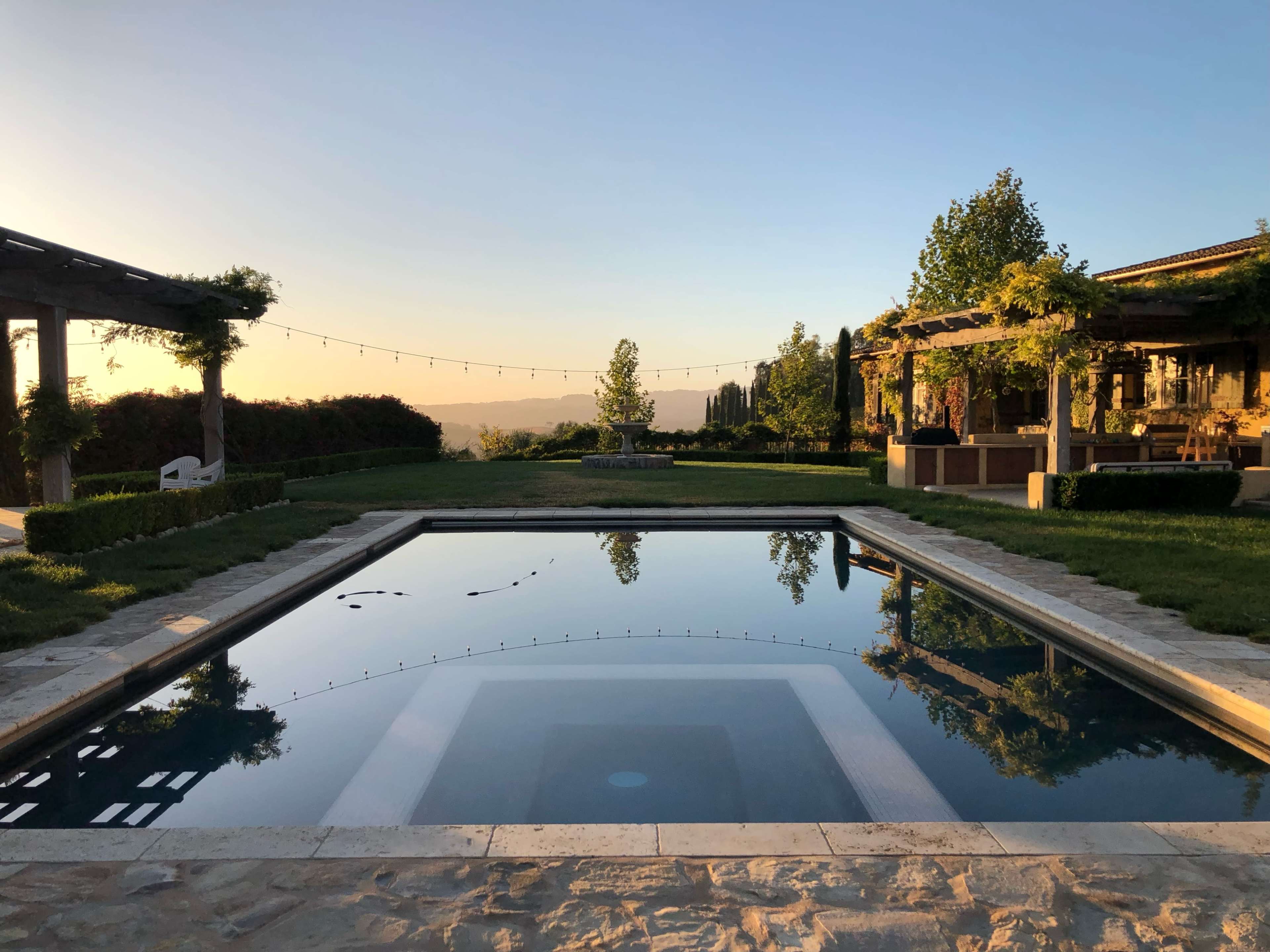 A rectangular pool reflects the surrounding landscape and structures during sunset, with string lights hanging above the area.