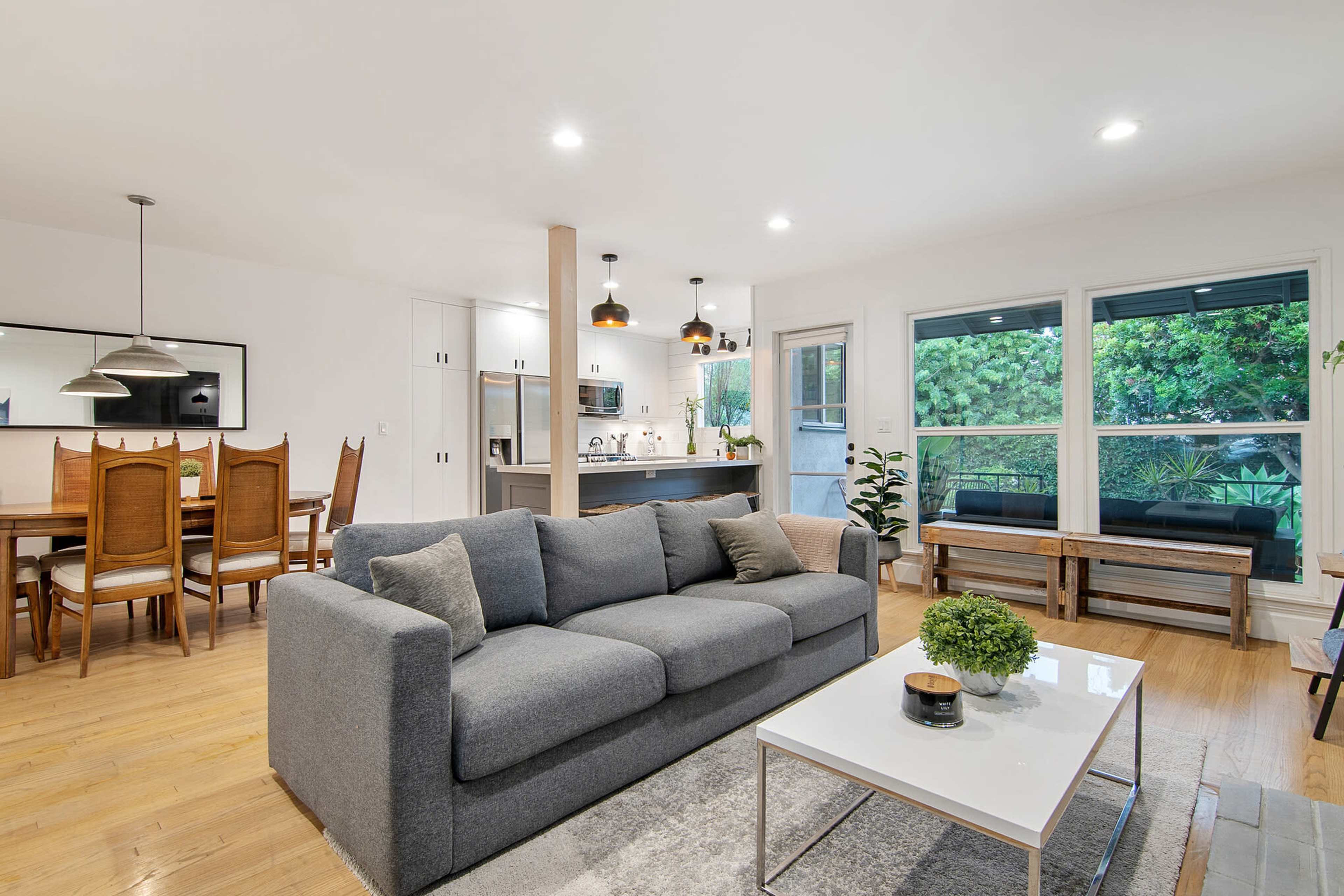 The image shows a modern living room with a grey sofa, a coffee table, and a dining area in the background featuring a wooden table and chairs.
