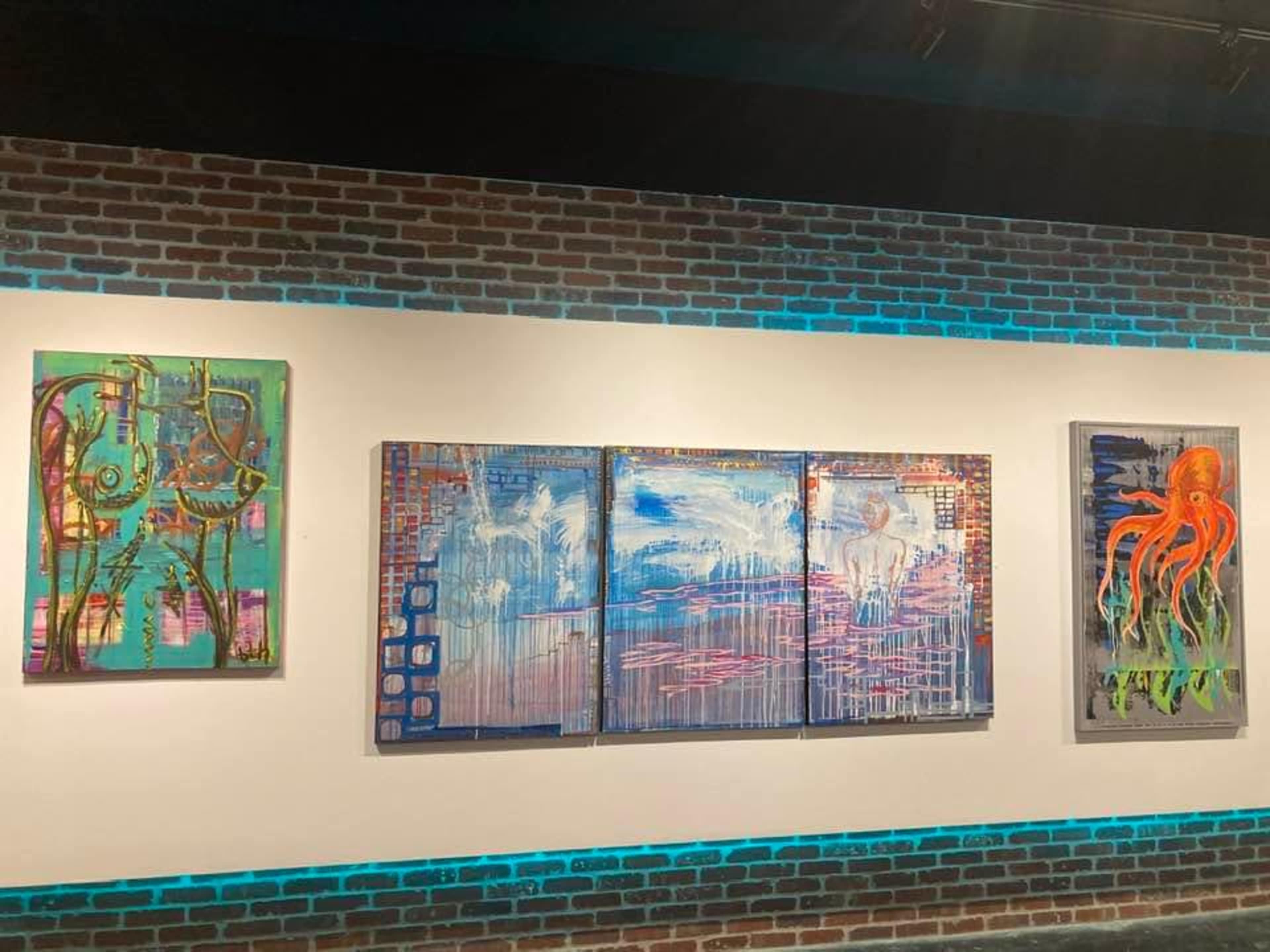 A gallery wall featuring five colorful abstract paintings, including an octopus, displayed against a brick background with blue lighting.