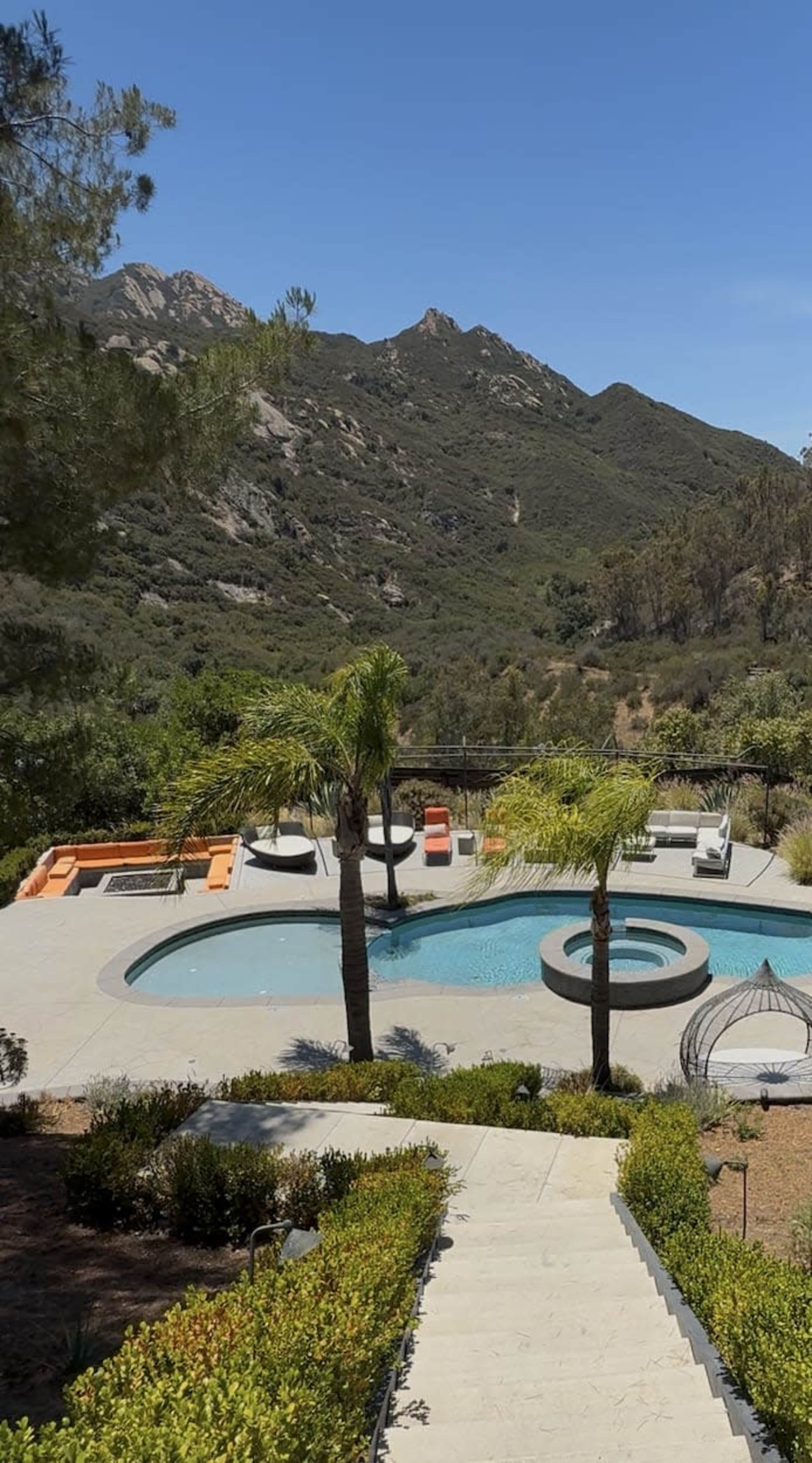 The image shows a modern pool area surrounded by greenery, with a backdrop of mountains under a clear blue sky.
