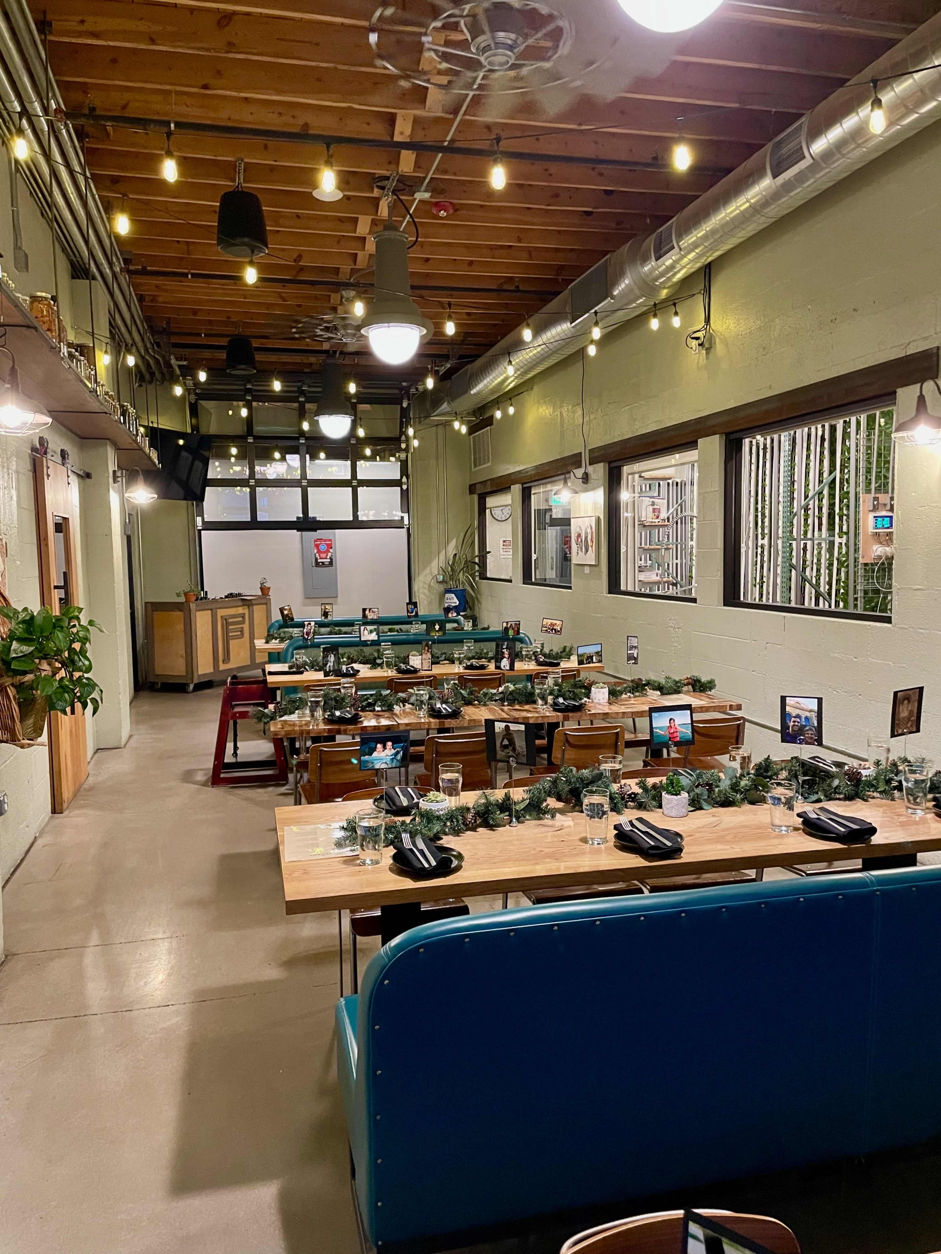 A restaurant interior with tables set for dining, decorated with greenery along the center, and vintage photos hanging on the walls under string lights.