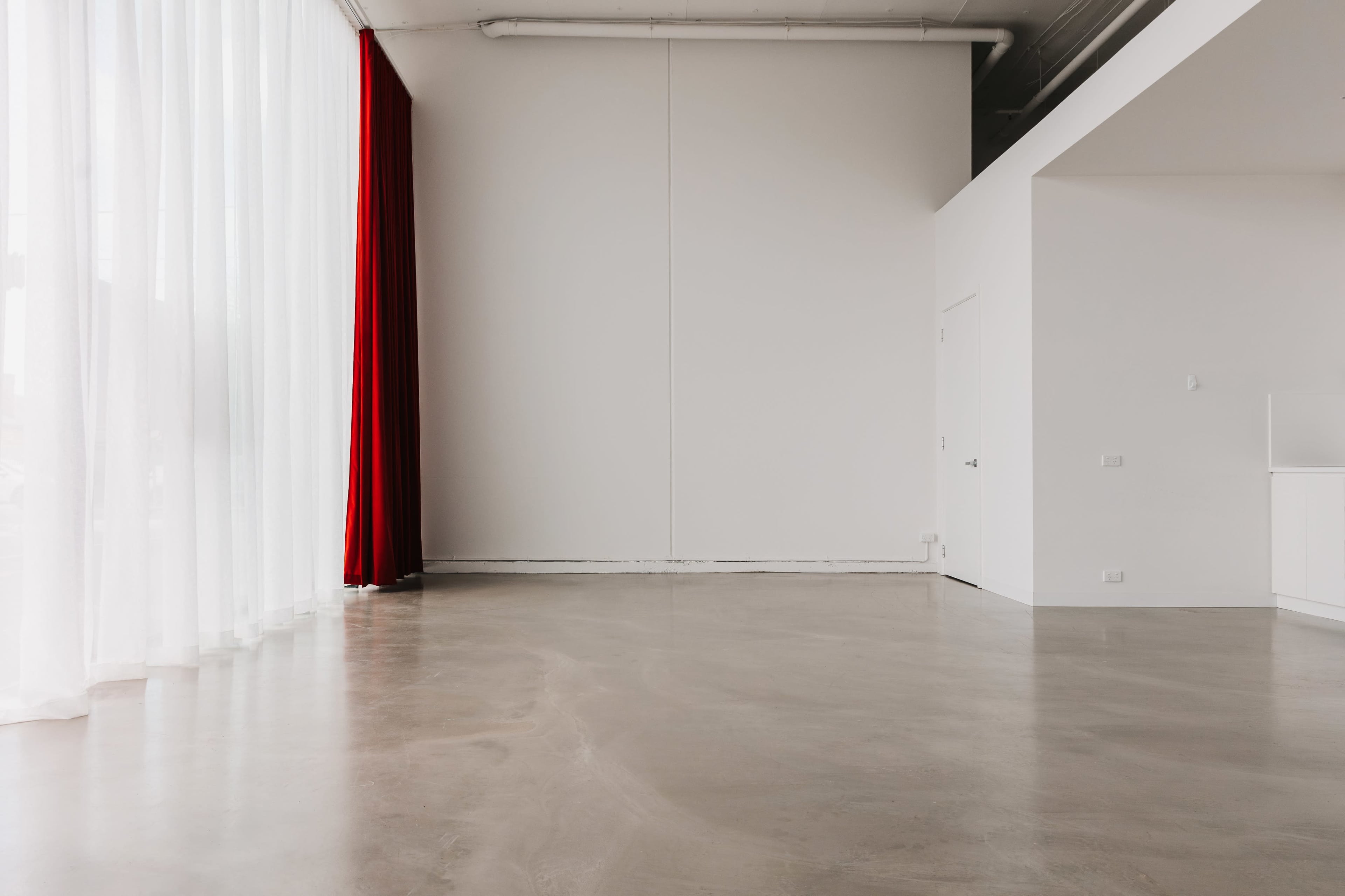 A spacious interior features polished concrete flooring, white walls, and a large window draped with sheer curtains and a red curtain.