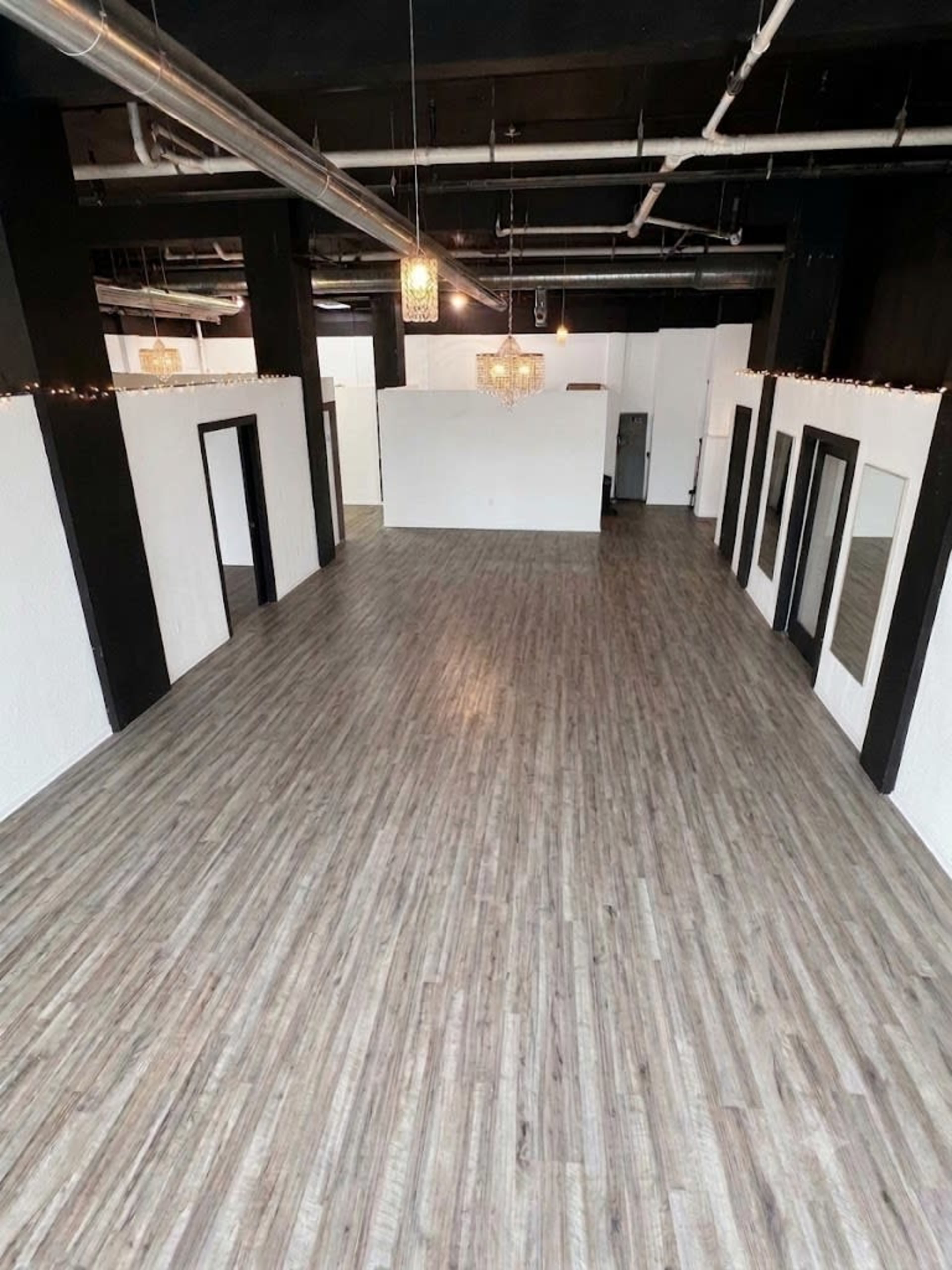 The image shows a spacious, empty interior with wooden flooring, black pillars, and white walls, featuring several doorways and mirrors.