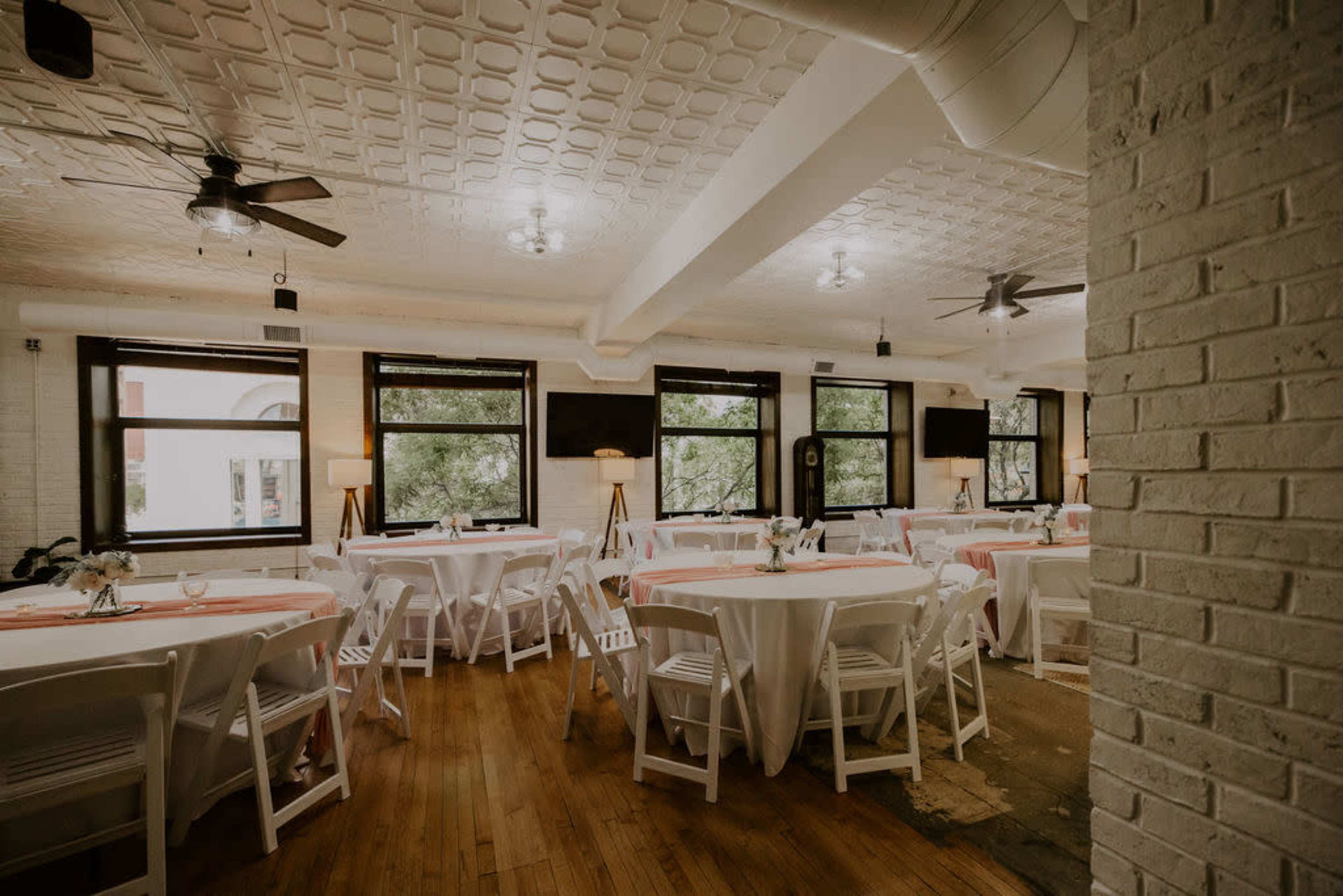 A spacious event room features round tables with white tablecloths and pink accents, arranged in a clean layout under ceiling fans and bright windows.