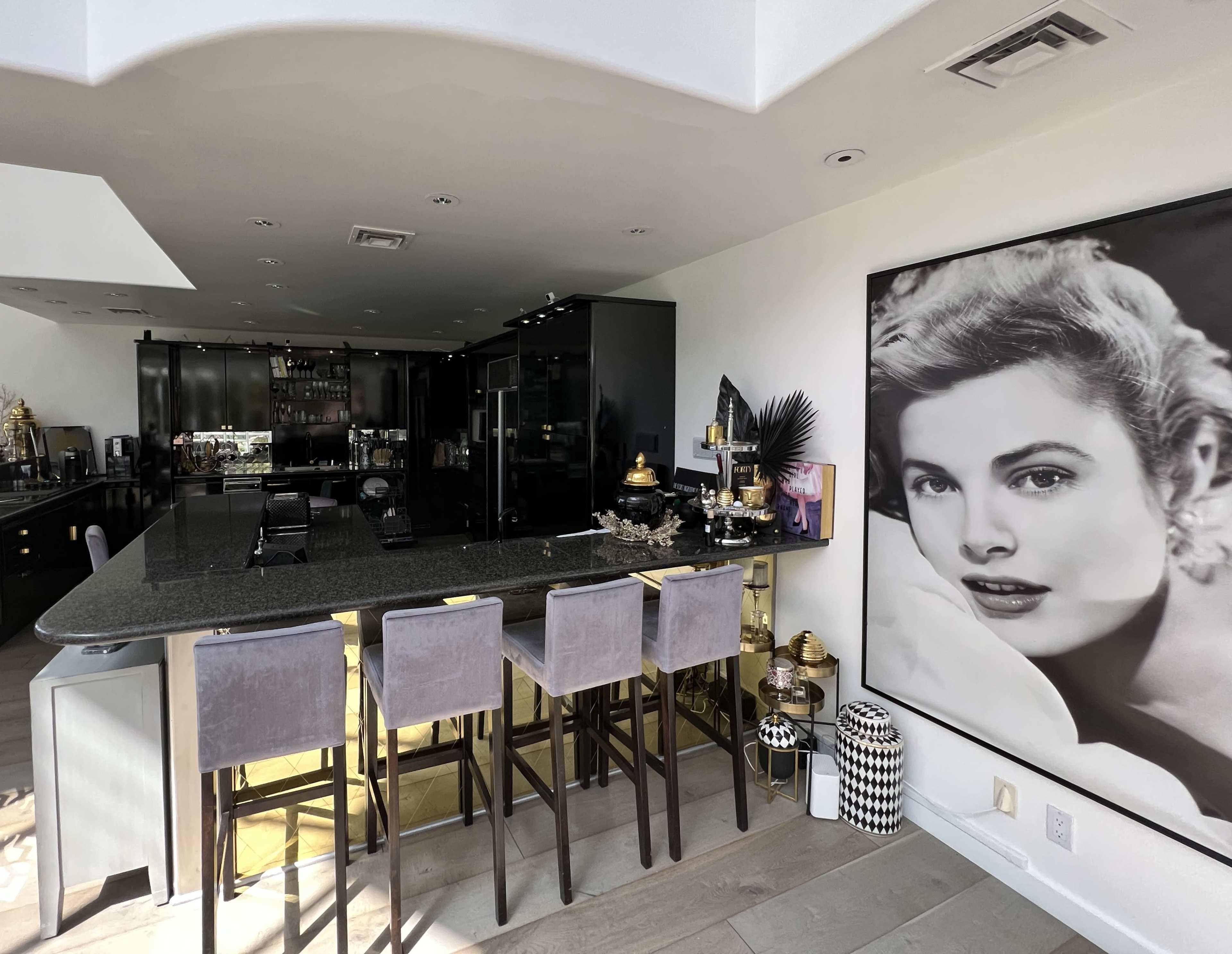 A modern kitchen with sleek black cabinetry and a bar area featuring four gray stools, alongside a large black-and-white portrait on the wall.
