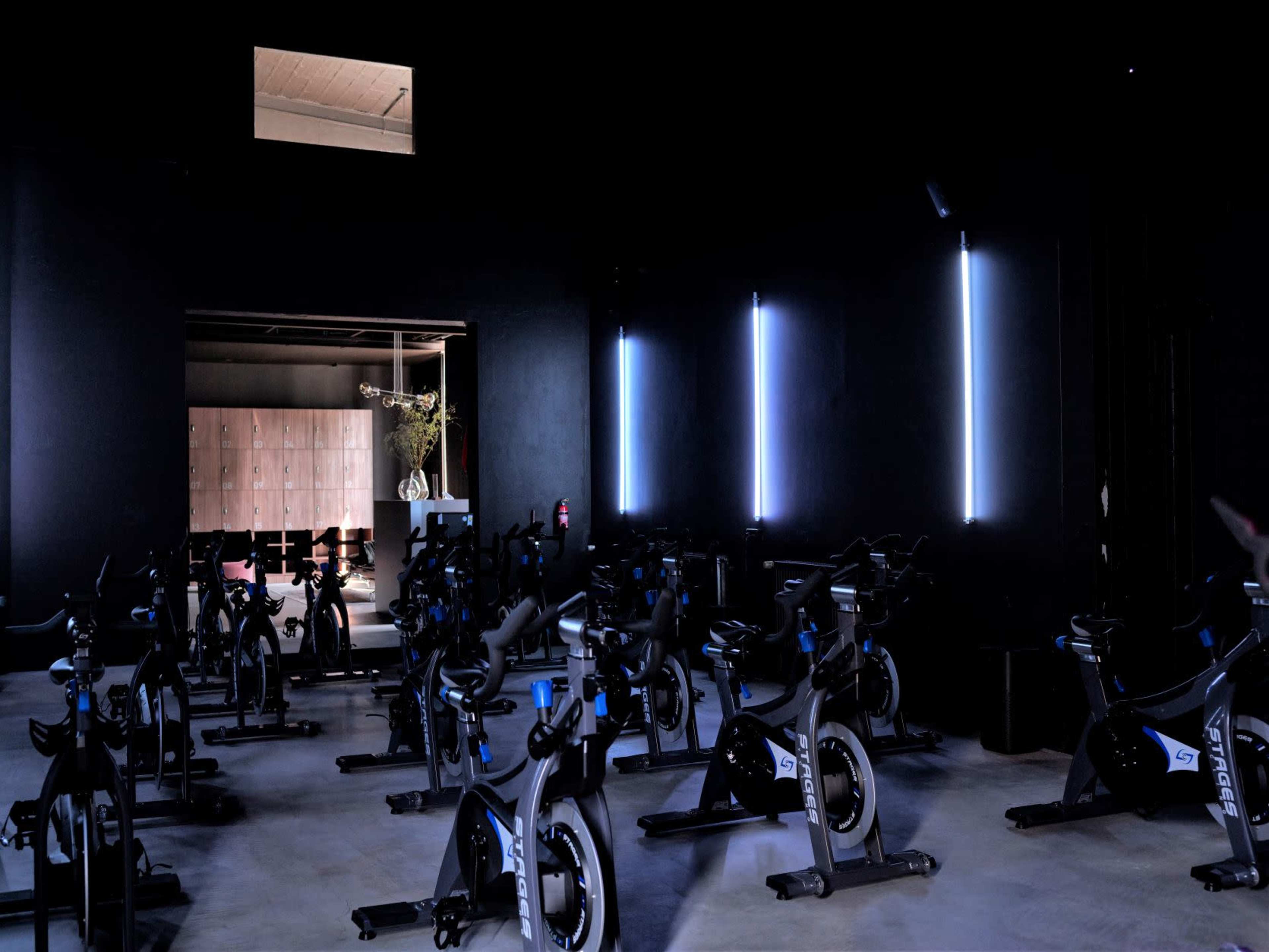 A dark indoor cycling studio features rows of stationary bikes illuminated by vertical strips of blue light.