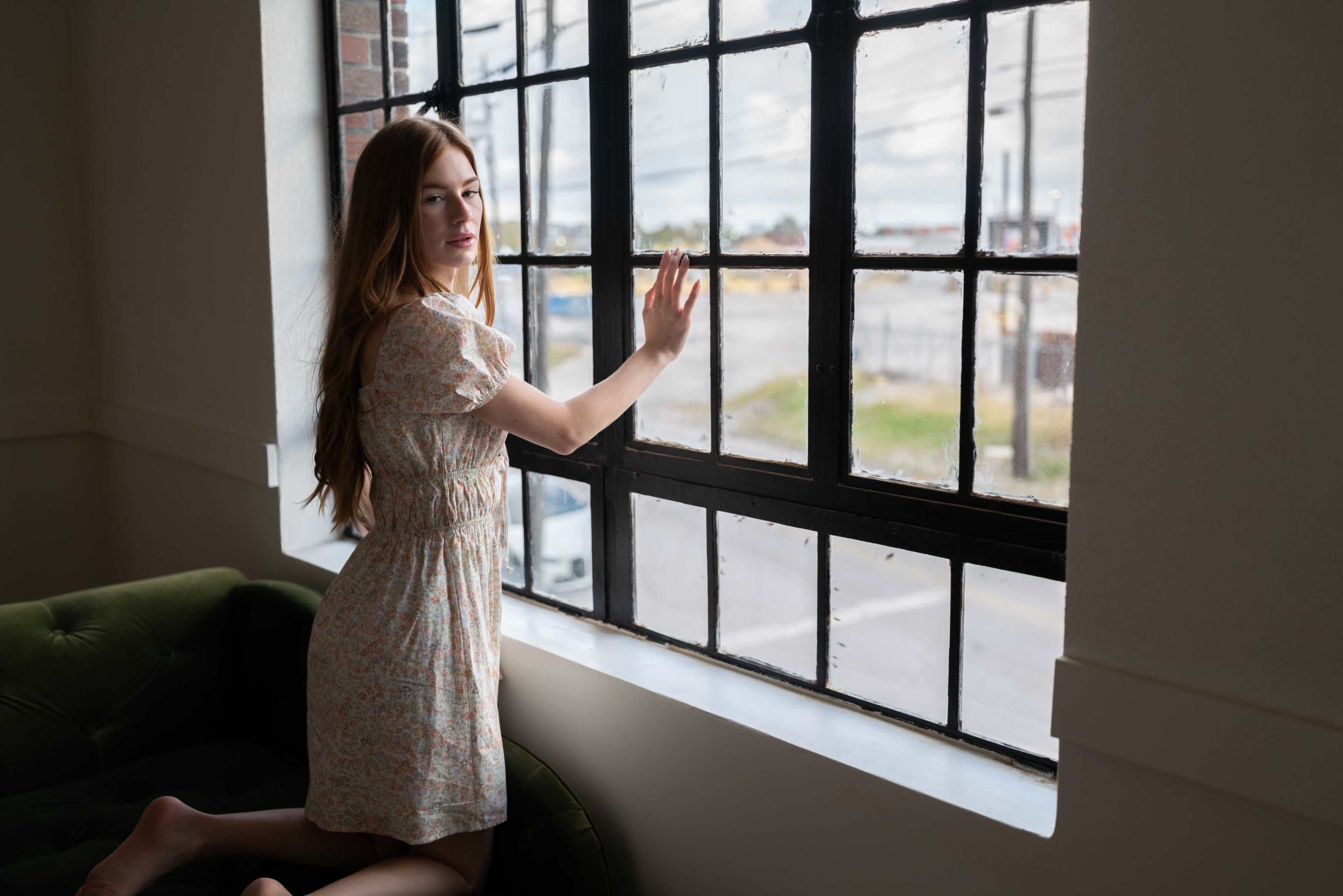 A young woman in a floral dress leans against a large window, looking outside while resting her hand on the glass.
