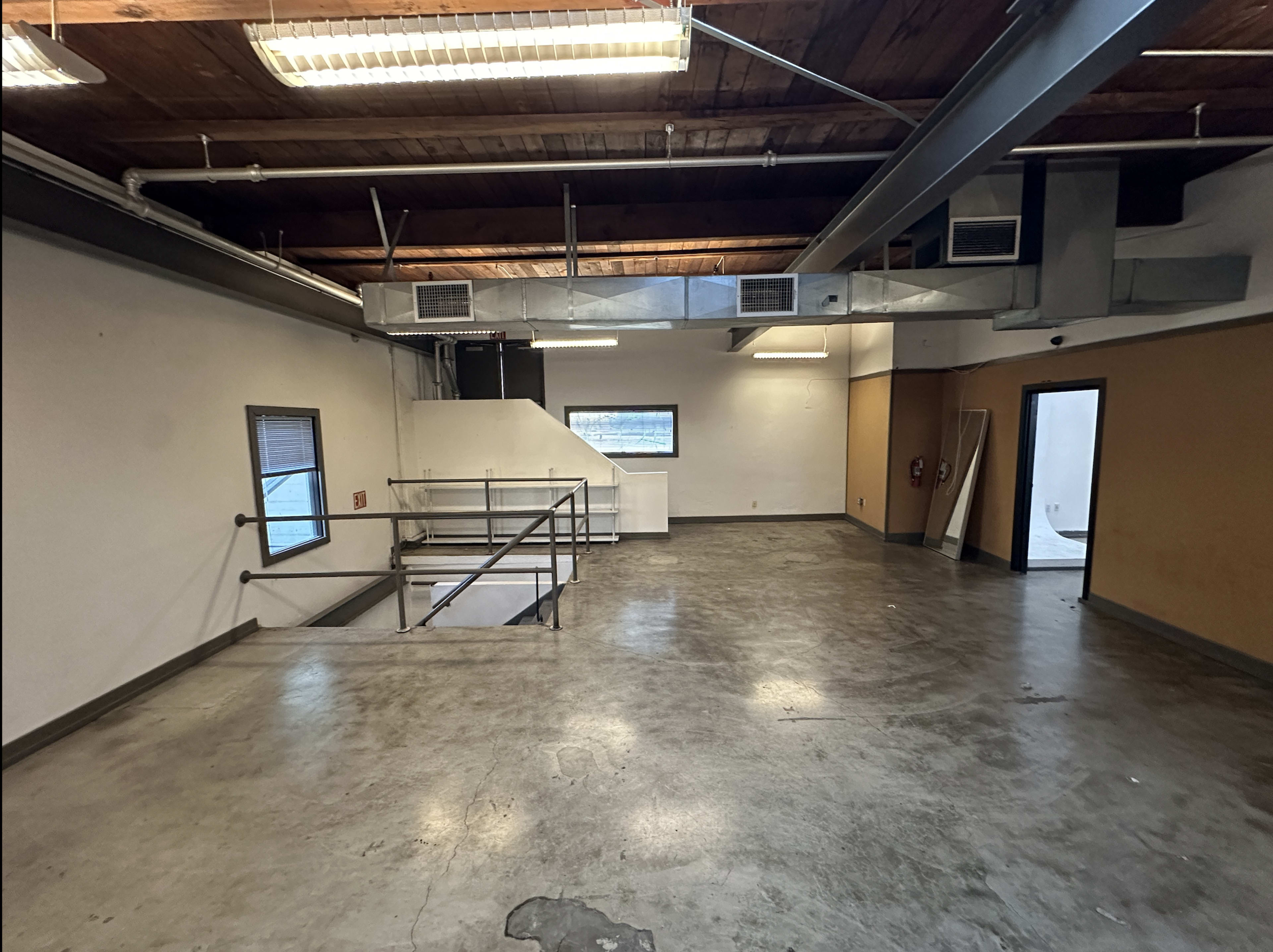 The image shows a spacious, unfinished interior with high ceilings, exposed beams, and concrete floors.