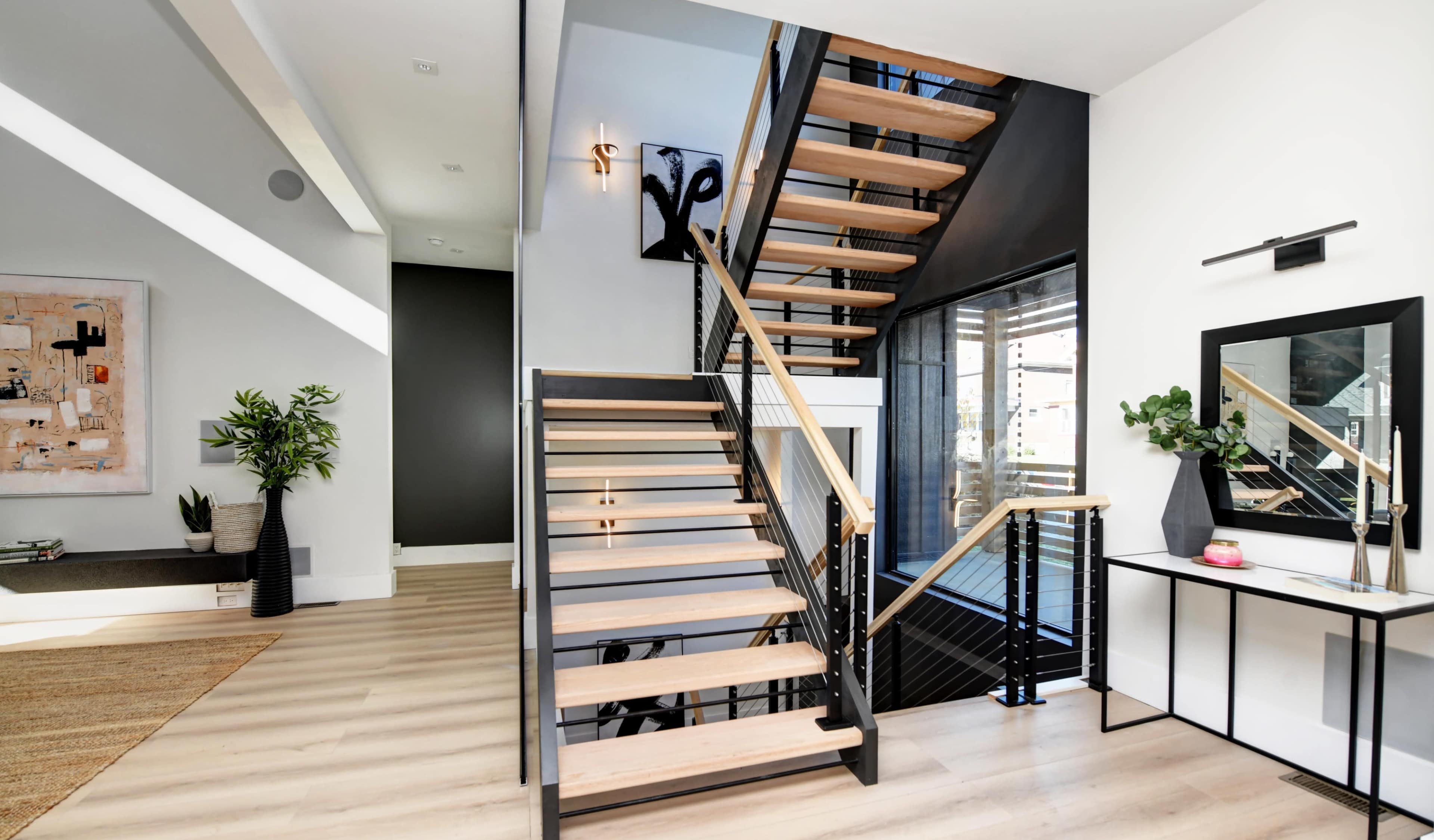A modern interior space featuring a wooden staircase with black railings, surrounded by minimalist decor and large windows.
