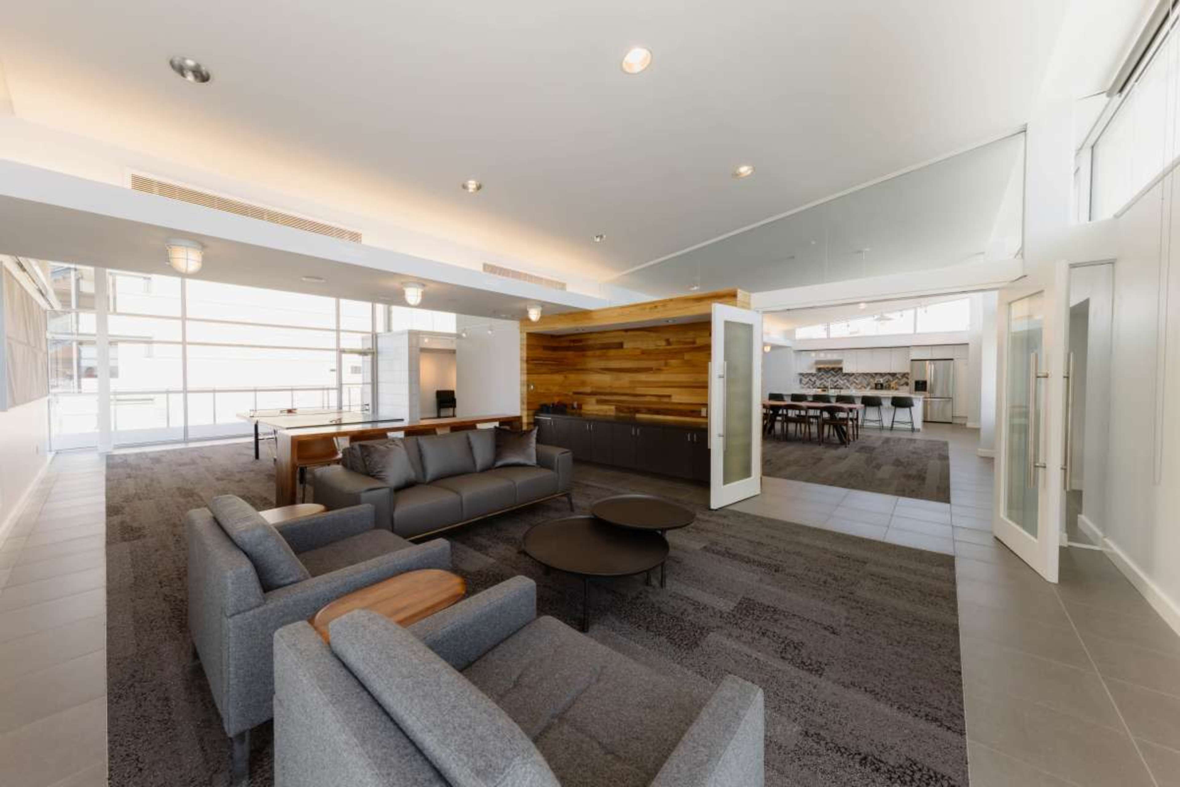 The image shows a modern lounge area with gray sofas, a circular table, and a wood-paneled feature wall, leading into a kitchen and dining space.