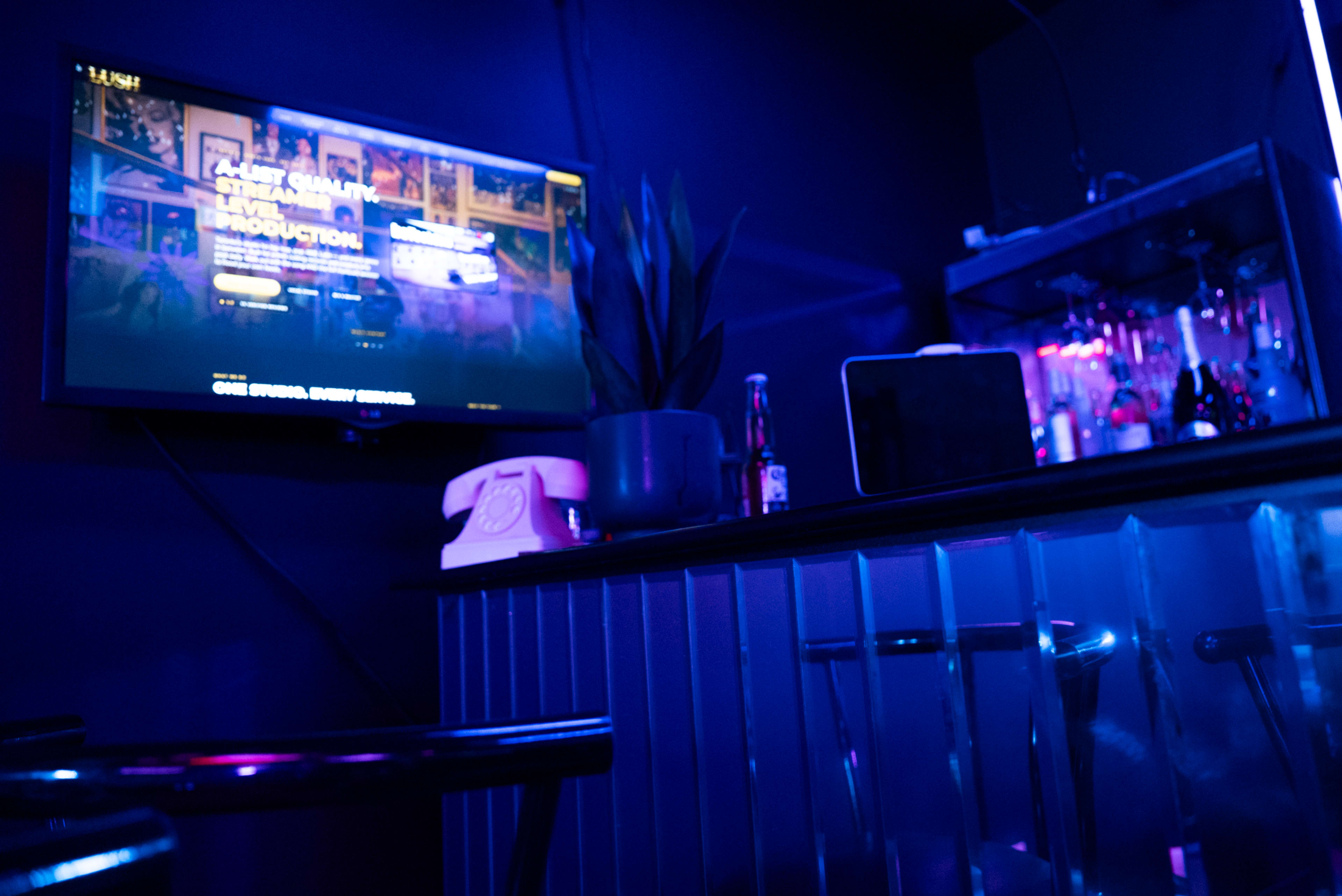 A dimly lit bar area features a television displaying a promotional screen, a potted plant, and a counter with glassware and a vintage telephone.