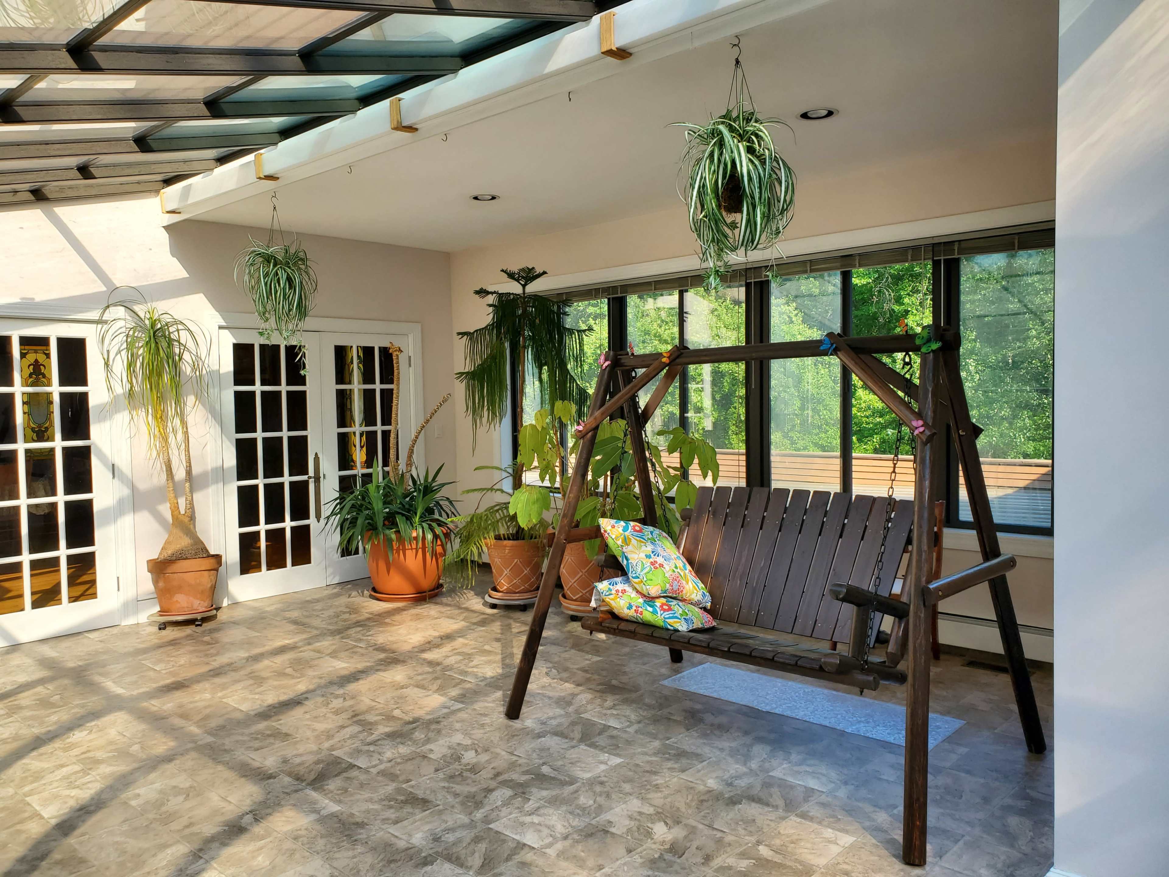A wooden swing is positioned in a sunlit room surrounded by large windows and several potted plants.