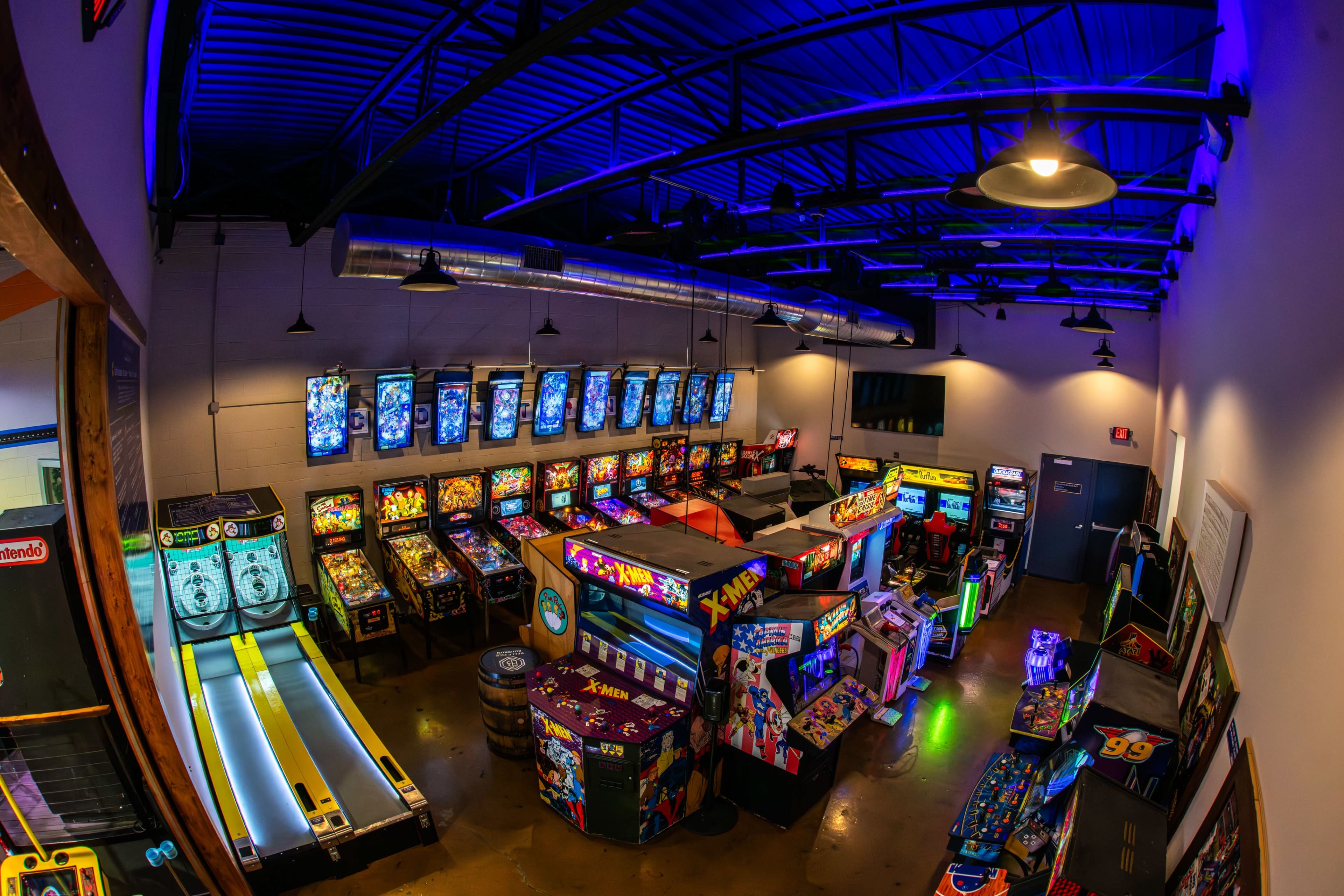 The image shows an arcade filled with various pinball machines and video games under blue lighting.