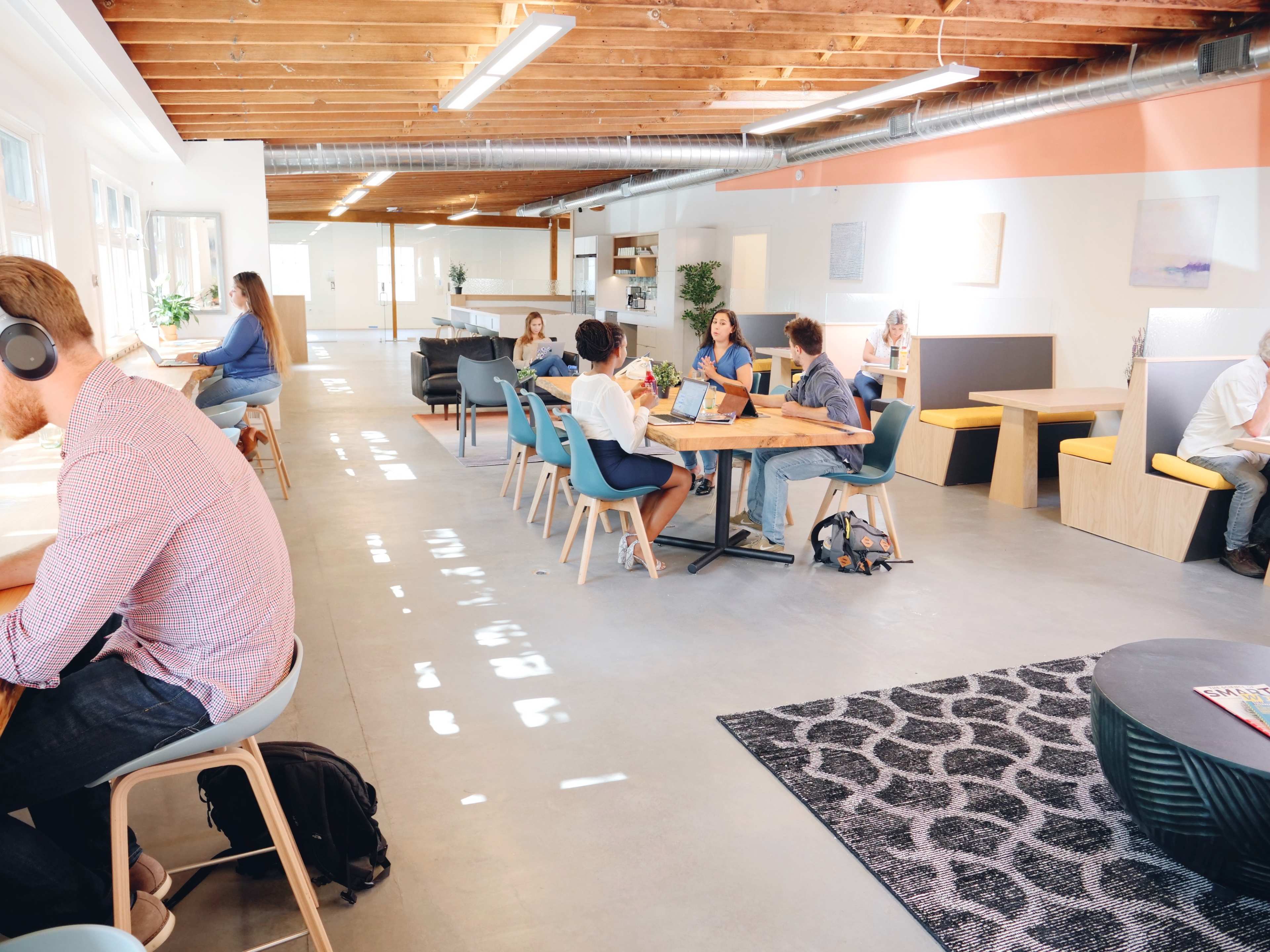 A modern workspace features people collaborating at tables, with a mix of seating arrangements and natural light flooding the area.