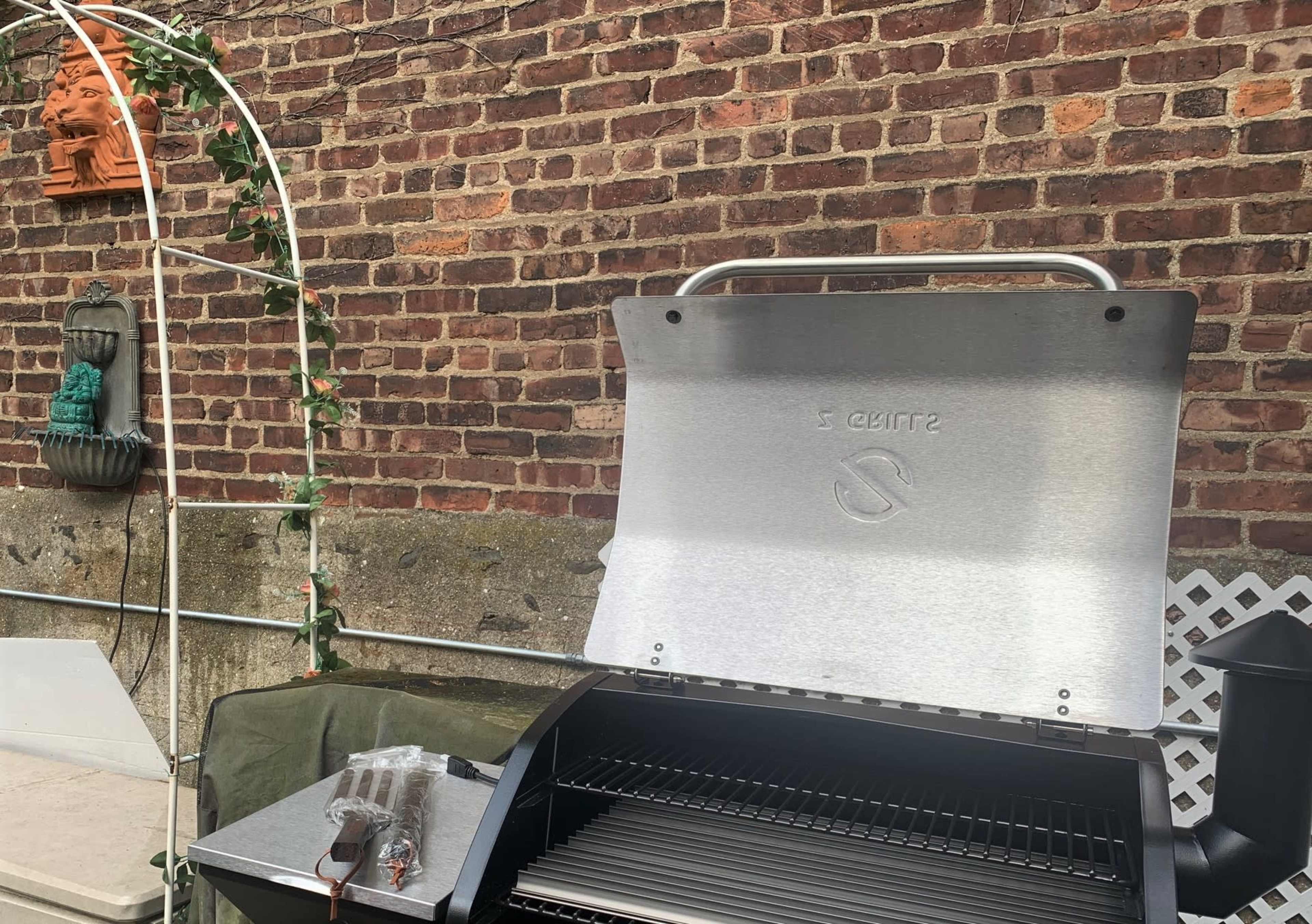 The image shows a stainless steel grill with its lid open, set against a brick wall and surrounded by decorative elements like an arch and greenery.