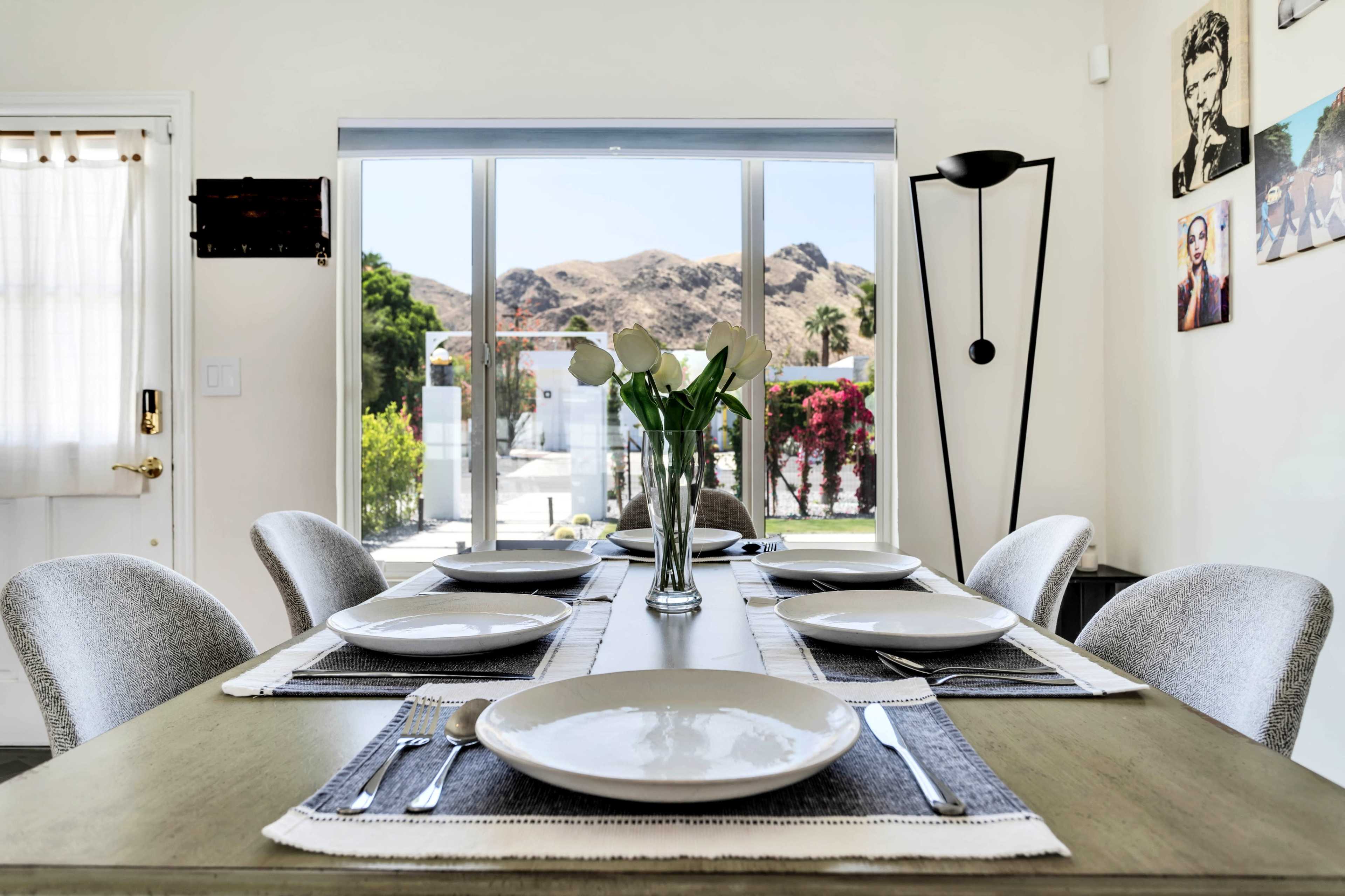 A dining table is set with plates and cutlery, and a view of mountains and greenery is visible through a large sliding glass door.