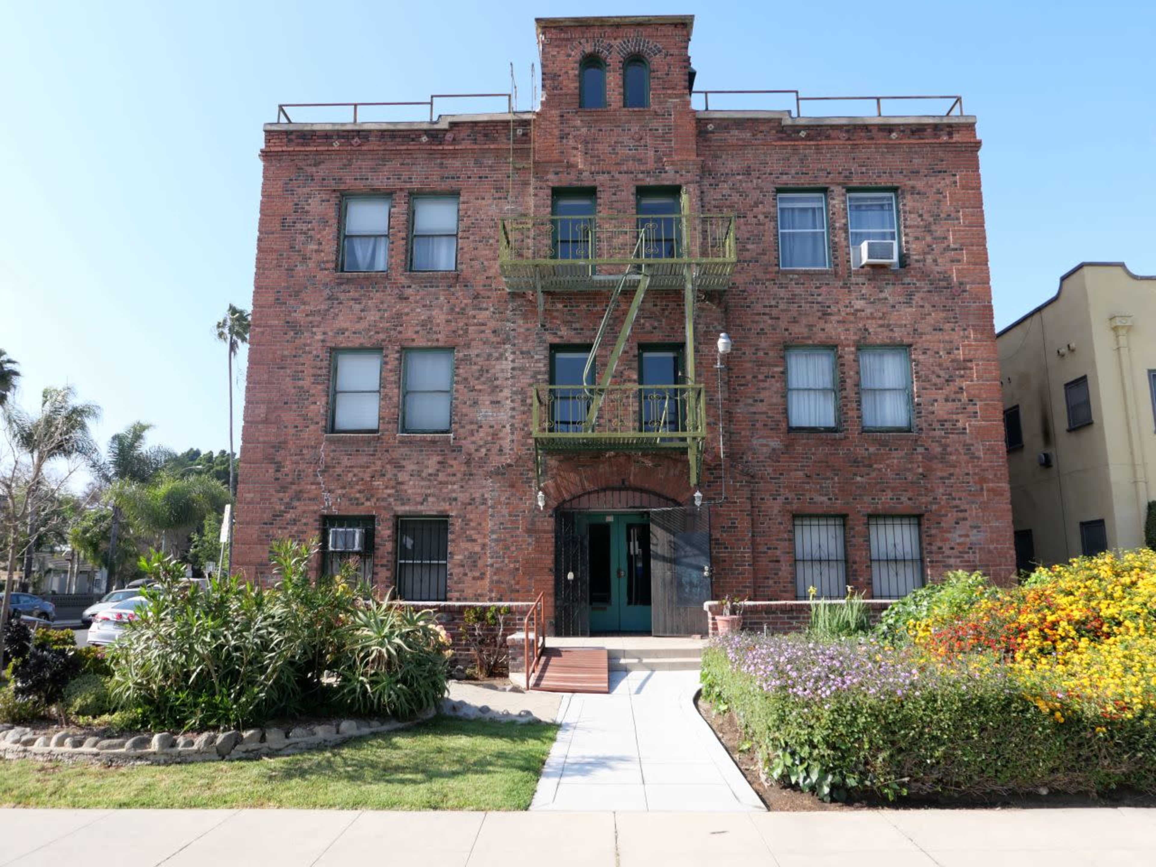 The image shows a red brick apartment building with green balconies and a staircase, surrounded by landscaped gardens and flowers.