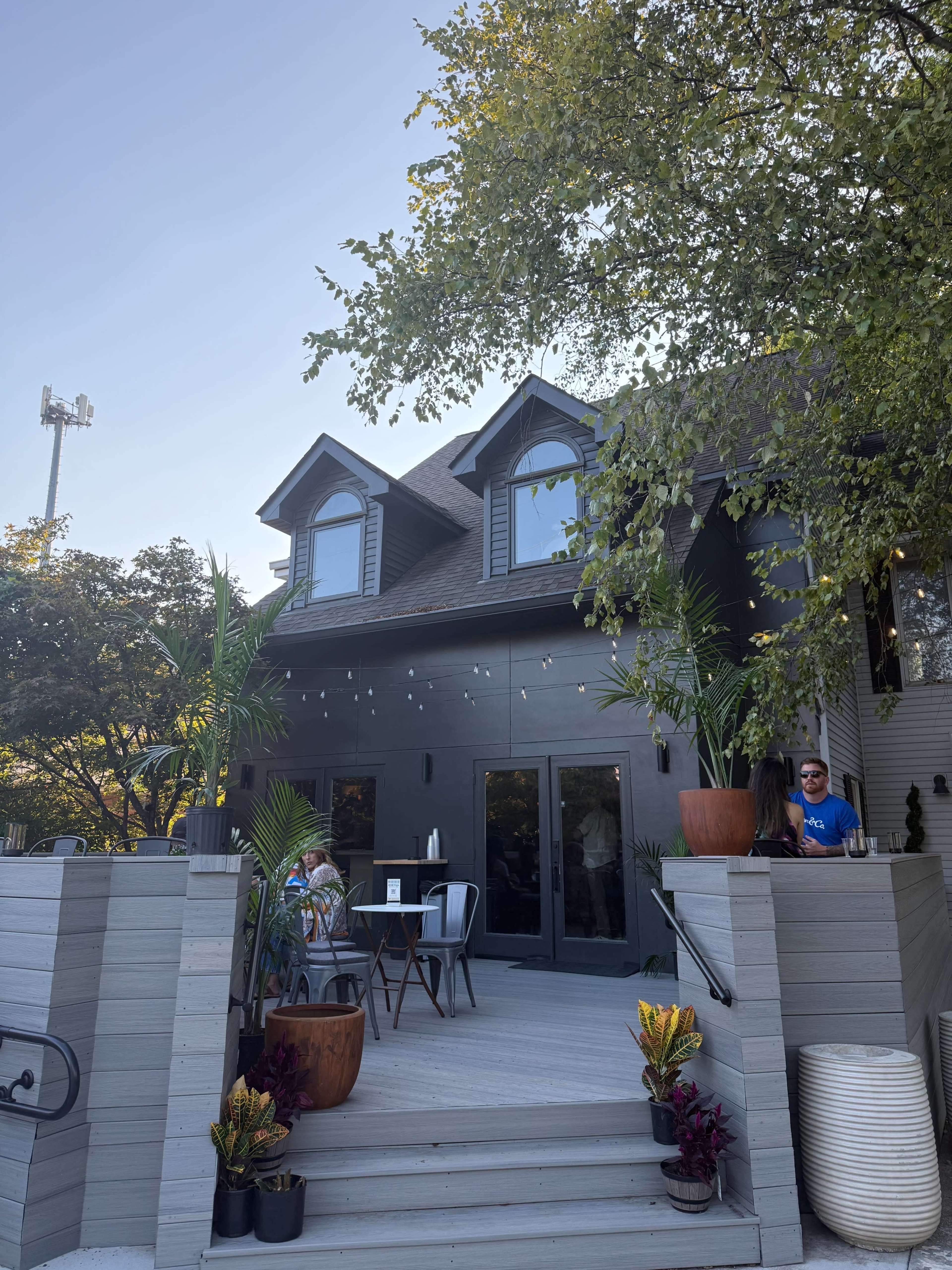 A two-story house with a dark exterior features a spacious deck adorned with plants and outdoor seating.