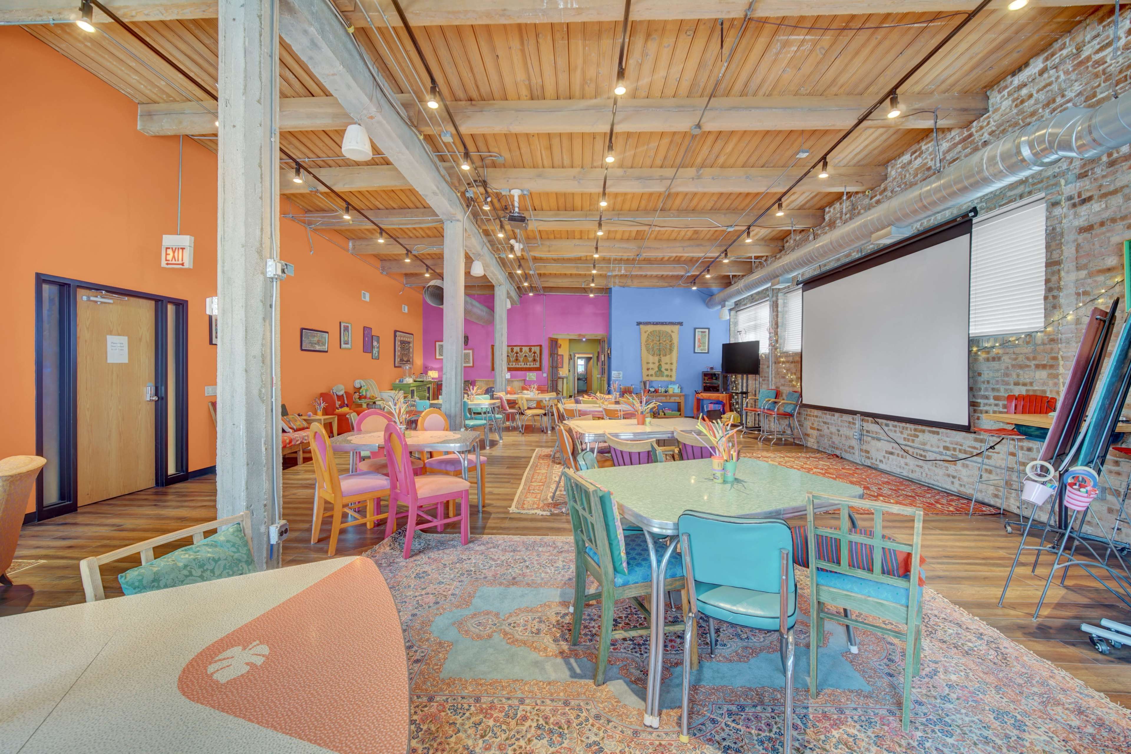 A spacious communal room with colorful furniture, exposed wooden beams, and a large projection screen against a brick wall.