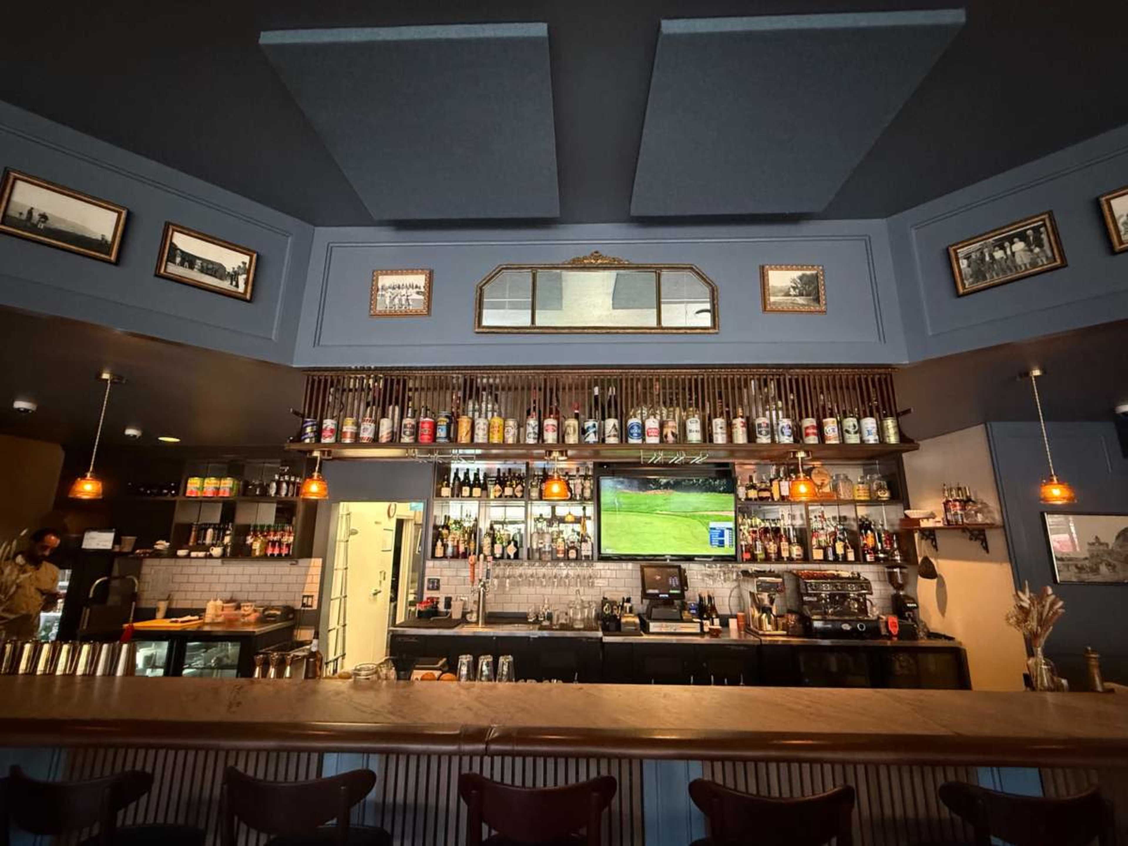 The image shows the interior of a bar with a well-stocked liquor shelf, a large screen displaying a sports game, and framed photographs on the walls.