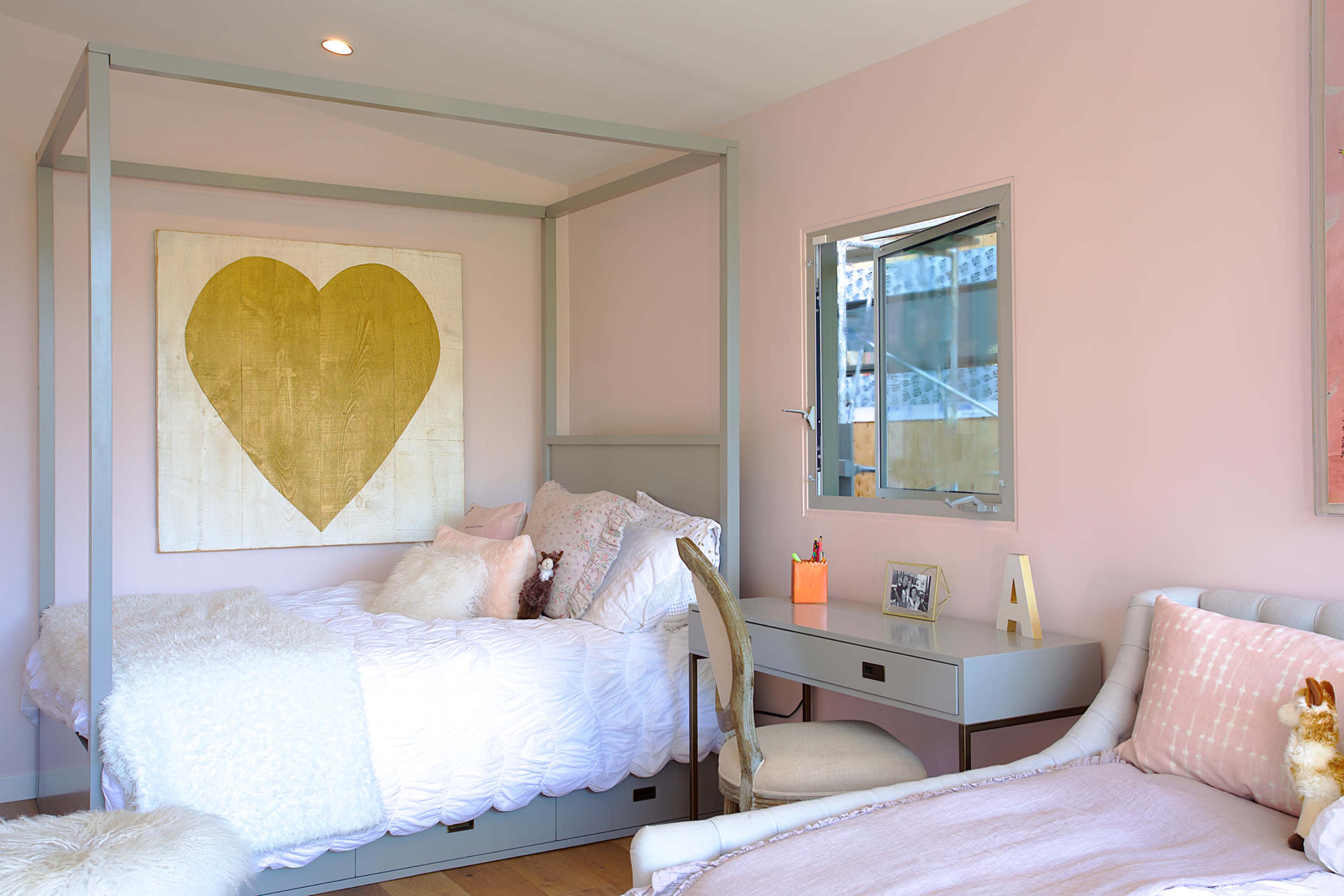A bedroom with a four-poster bed, a heart-shaped gold artwork on the wall, and a desk with a chair beside a window.